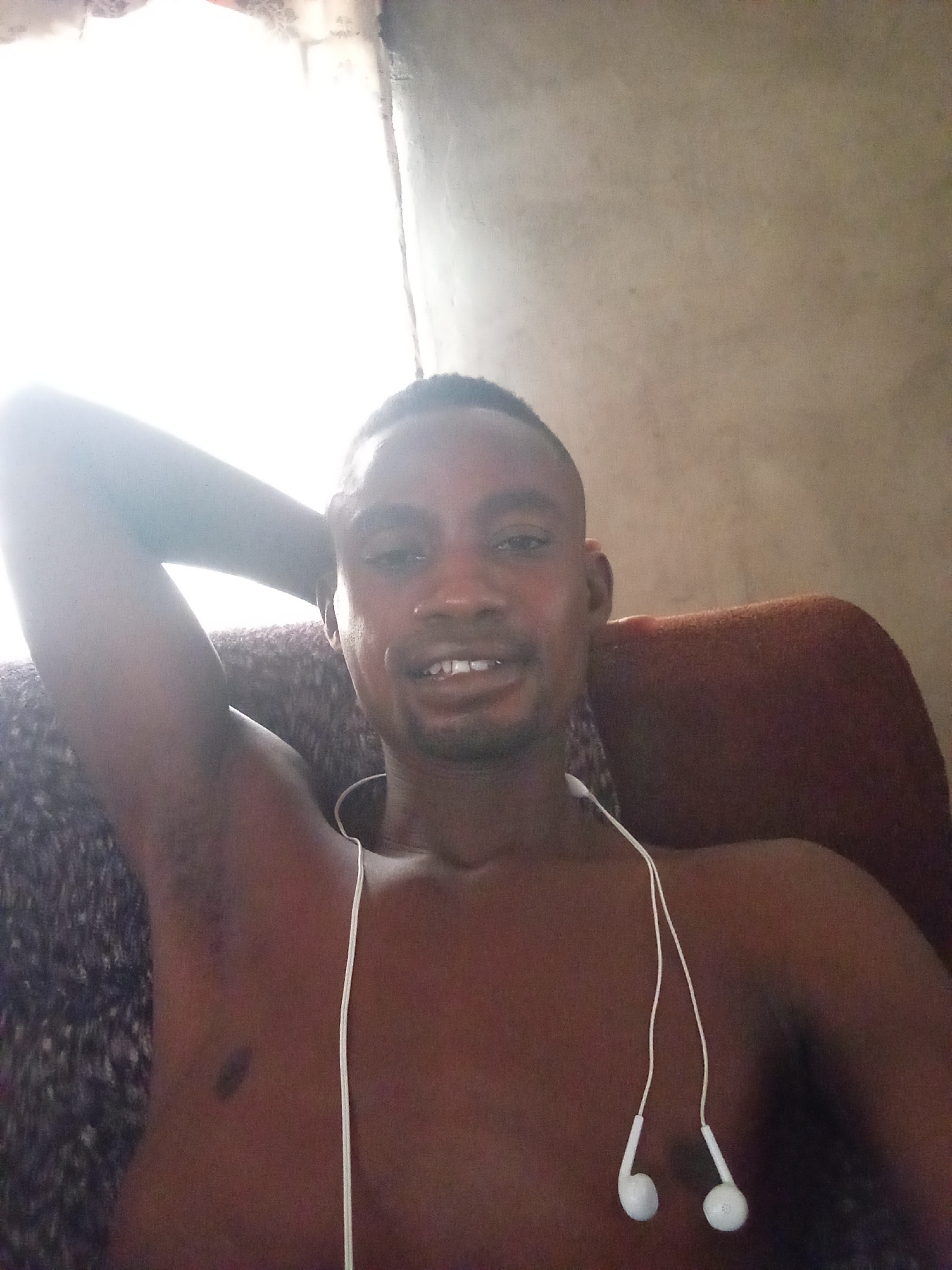 A shirtless man with short-cropped hair is lying on a couch, with his right arm raised and resting behind his head. He has earbuds hanging around his neck. The background appears to be an interior space with a window providing natural light. The location and city of Adonkolo, Nigeria, are not discernible from the image.