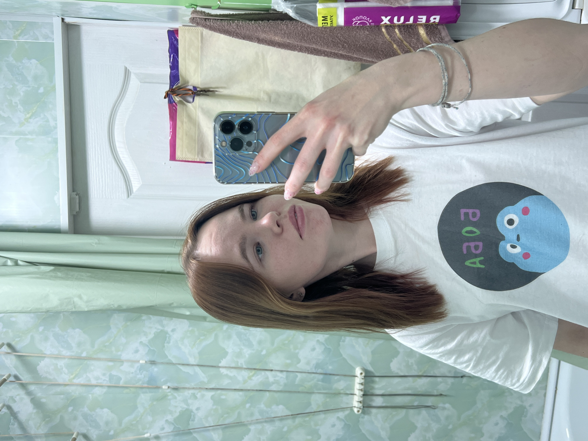 A woman with reddish-brown hair and fair skin takes a selfie in what appears to be a bathroom. She is holding a smartphone in her left hand, showcasing a clear case with a blue design and multiple camera lenses. She is wearing a white T-shirt with a circular graphic featuring a light blue character and the letters "Aaoa" in different colors.
The bathroom has tiled walls in a light green marble pattern. A white door is visible in the background, along with a towel rack and various bathroom items on a shelf above. The location appears to be in a residential setting, and the description aligns with a possible location in the city of Chaikovsky, Russia, given the common residential bathroom styles.