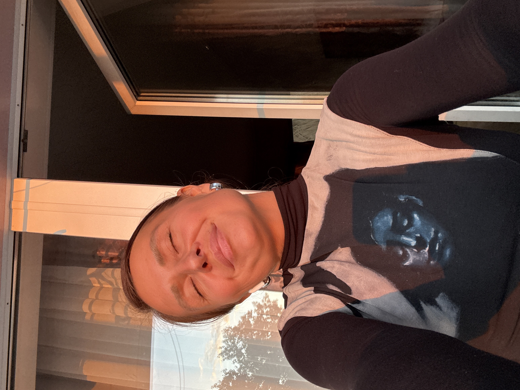 A woman is shown in an indoor setting, angled to the left. She has fair skin, closed eyes, and is sticking out her tongue. She's wearing a black turtleneck under a patterned shirt featuring a prominent dark figure. She is positioned near a window, which is partially open at the top. The scene is illuminated by a golden light, possibly from the setting sun. The location is Chaikovsky, Russia.