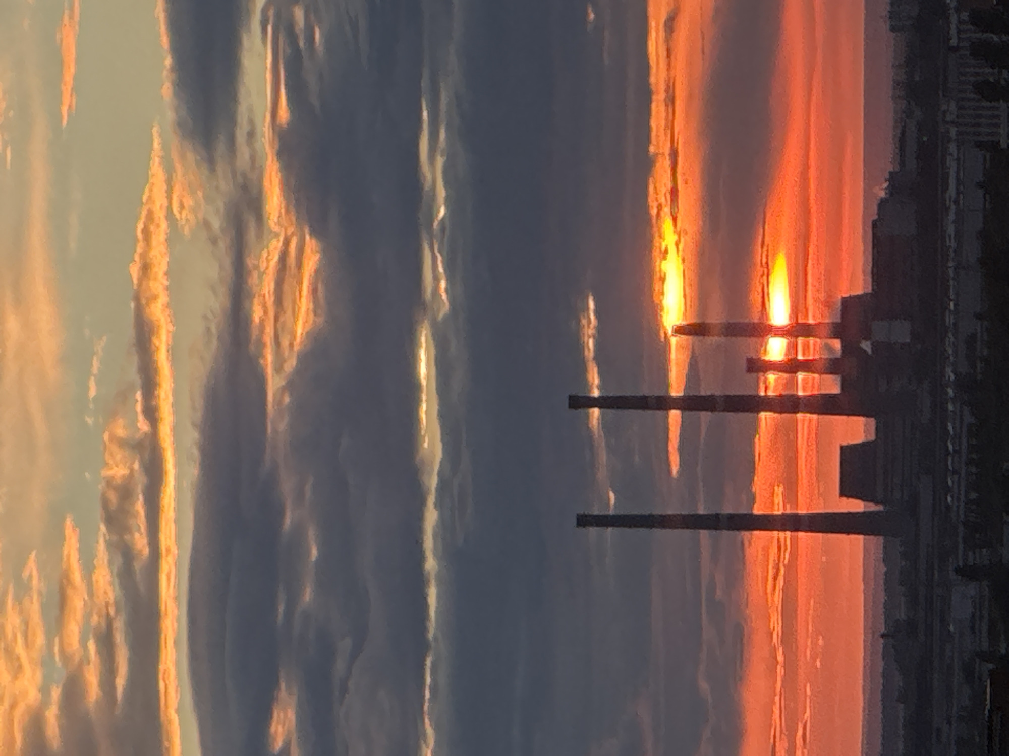 A vertical, medium shot captures a dramatic sunset over a city. The sky is filled with layers of clouds, the lower ones dark and tinged with gray, while the upper ones are illuminated by the setting sun in vibrant hues of orange and yellow. The sun itself is a bright, fiery orange orb peeking through a horizontal band of clouds. In the foreground, silhouetted against the sunset, stand several tall, cylindrical smokestacks of an industrial facility. The structures appear to be part of a power plant or factory complex. Below the smokestacks, a dark, indistinct urban landscape stretches towards the horizon. The overall impression is one of industrial power set against the natural beauty of a sunset.