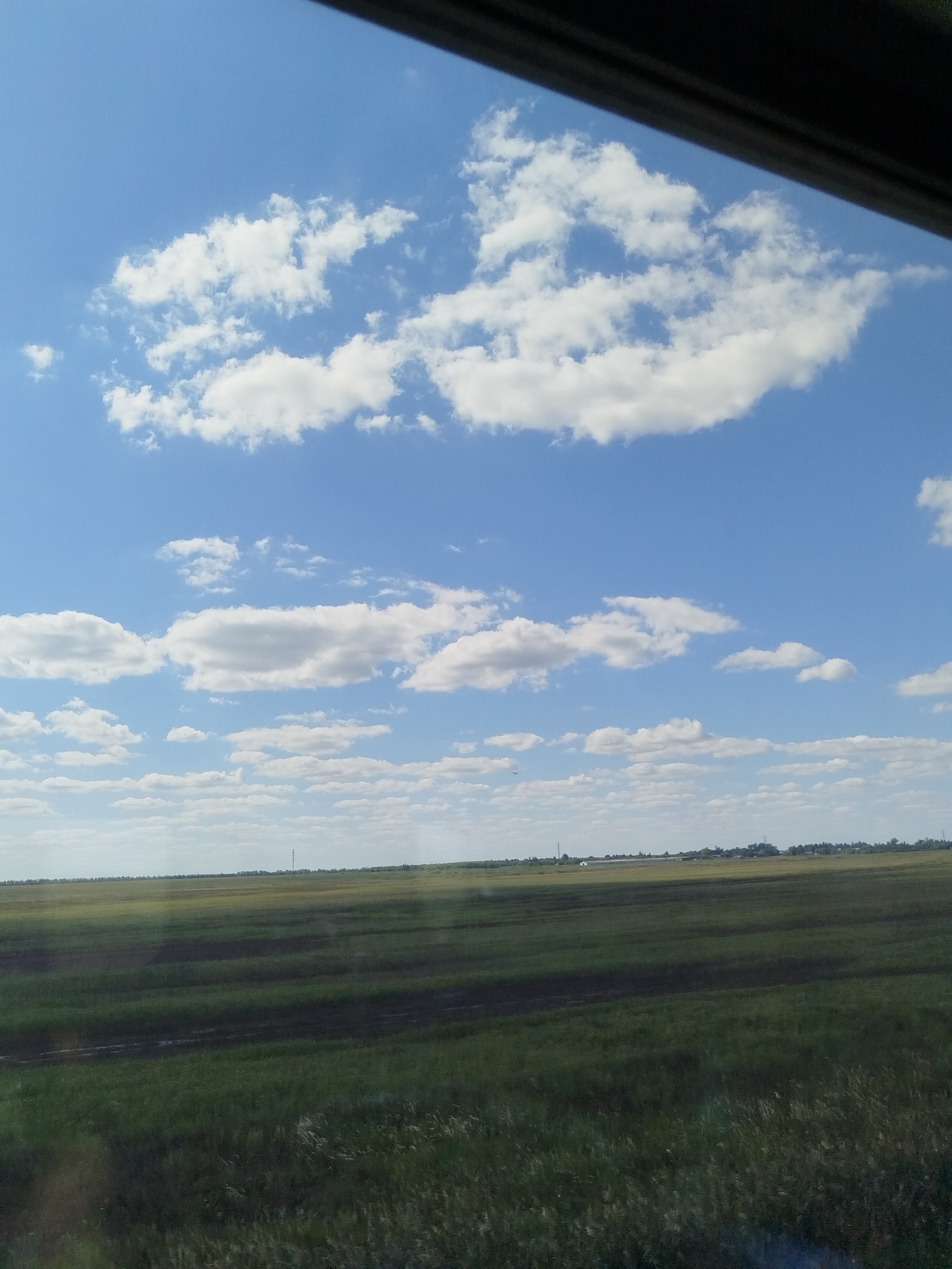 The image depicts a wide, flat landscape under a bright blue sky dotted with fluffy white clouds. In the foreground, a field of green grass is visible, with darker, tilled or unplanted strips running through it. In the distance, a faint line of trees marks the horizon, and several small structures, possibly buildings or farmhouses, can be discerned. A tall, slender tower, likely a communication or observation tower, stands prominently against the sky. The scene appears to be a rural or agricultural area, characteristic of the landscape found in and around Korneevka, Russia. The perspective suggests the image was taken from a moving vehicle, possibly a car or train, given the slight distortion and the dark frame at the top of the image.
