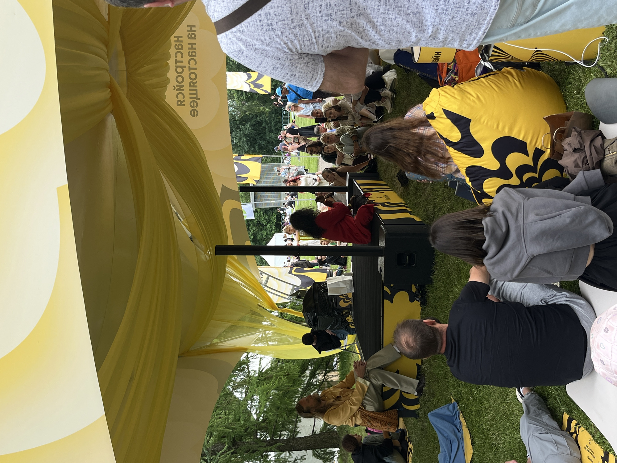 A ground-level outdoor shot shows a stage set up beneath a yellow canopy, with an audience of people seated on the grass, facing the stage. The scene is likely at an outdoor event, possibly a concert or festival, given the setup and number of attendees. The dominant colors are yellow, black, and green from the canopy, the stage decorations, and the grass.
The location is Moscow, Russia, as the Cyrillic script on the yellow decoration shows the city name (Москва). The crowd is diverse in age and attire, sitting comfortably on the ground, some using blankets or cushions. The weather appears pleasant, and the event is in progress. The people seem focused on the stage.
