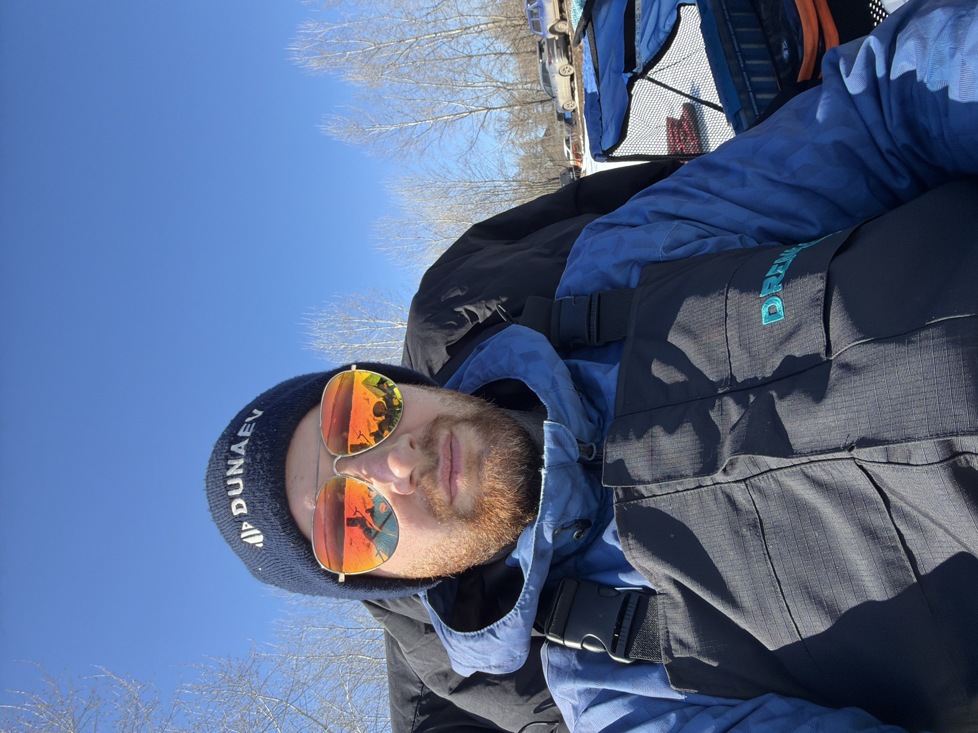 A selfie of a man with a beard wearing a blue beanie with the text "DUNAEV" and orange aviator sunglasses is shown. The sunglasses reflect a riverside location with ice fishing structures. The man is wearing a blue jacket under a black vest with "DREAMTEAM" visible in turquoise lettering. He is outdoors on a sunny day with bare trees and a blue sky. Cars are parked in the background. The location is Noviy, Russia.