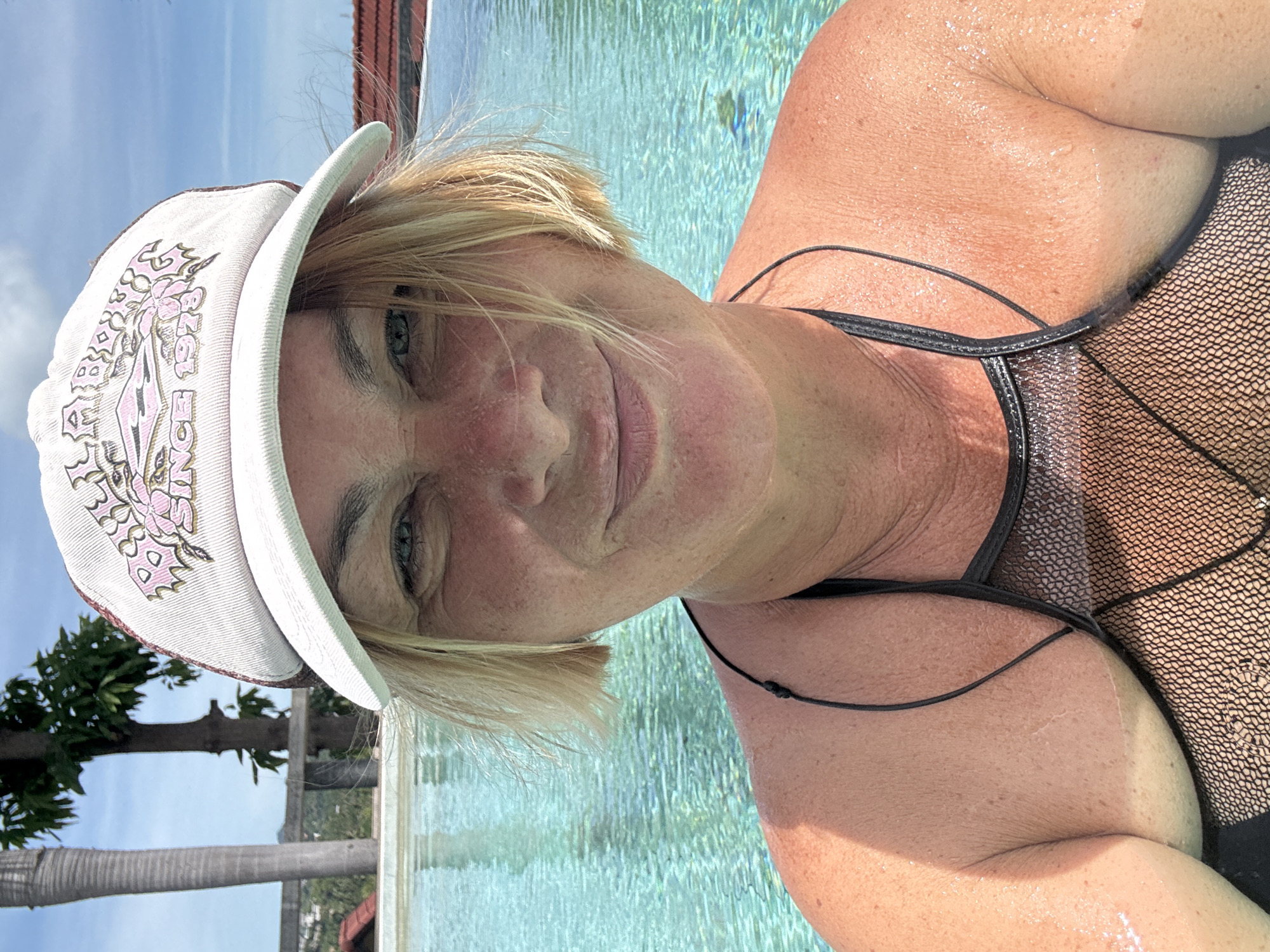 A middle-aged woman wearing a white cap with a pink design and a black swimsuit is in a swimming pool. The pool water appears turquoise, with the reflection of the blue sky visible. A tree and other foliage are visible on the left side of the frame. The location appears to be บ้านบางรักษ์, Thailand.
