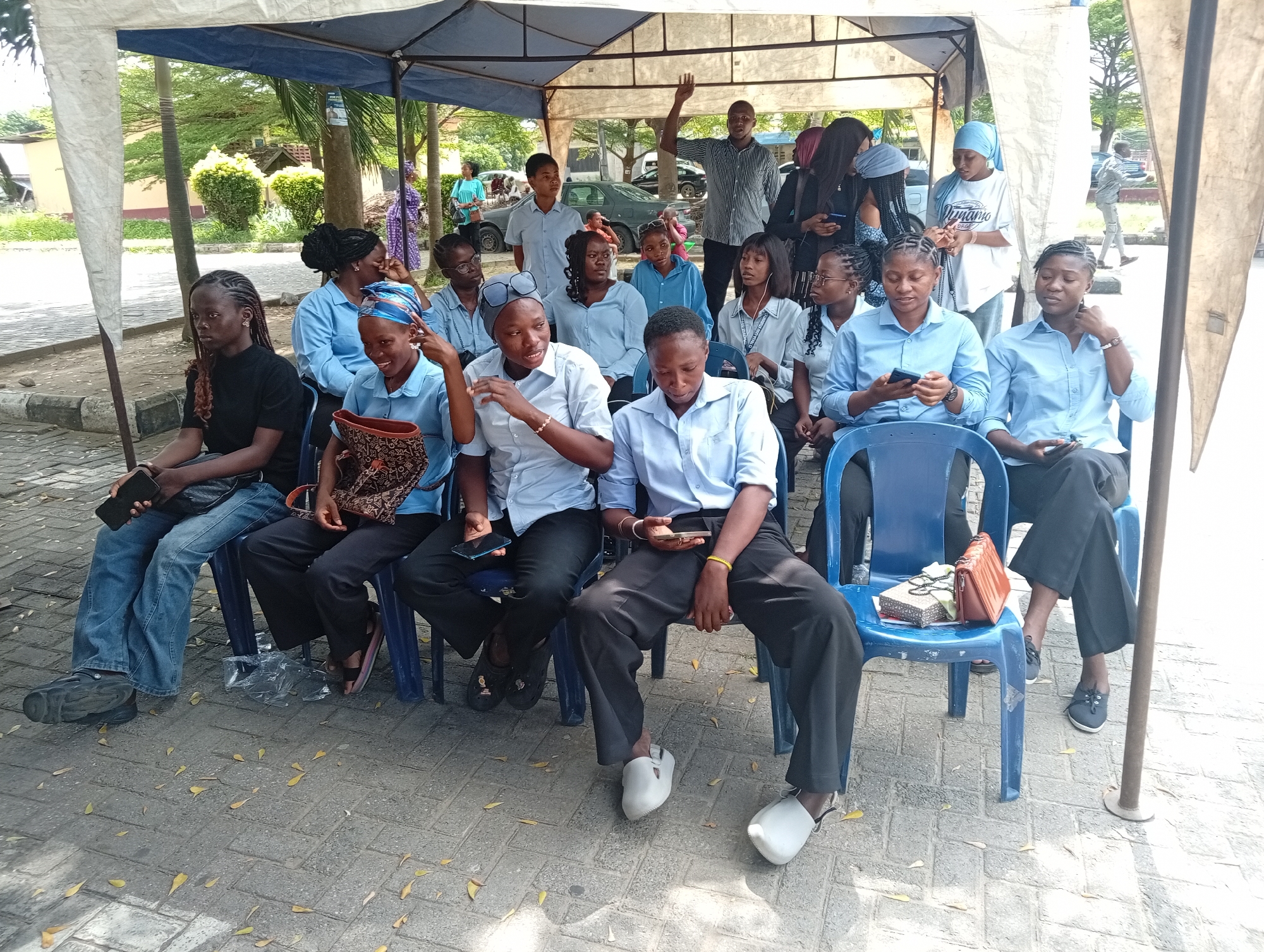 A group of approximately 15 young adults are seated on blue plastic chairs beneath a white canopy in Ijanikin, Nigeria. The setting appears to be an outdoor event or gathering.
Most individuals are wearing light blue collared shirts. Some are holding or using mobile phones. One man in the background is standing with his arm raised.
The ground is paved with interlocking concrete pavers, and scattered yellow leaves are visible. Trees and buildings are visible in the background.