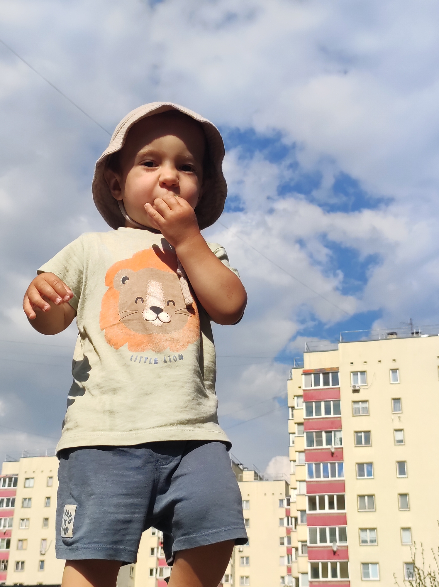 A toddler wearing a light-colored bucket hat, a light green t-shirt with a lion graphic, and dark blue shorts stands outdoors, facing the camera. The toddler is holding a finger to their mouth. In the background, there are two multi-story apartment buildings with yellow facades and red accents around the windows, under a sky with scattered clouds. The scene is likely in the city of Chaikovsky, Russia, as indicated by the architectural style of the buildings.