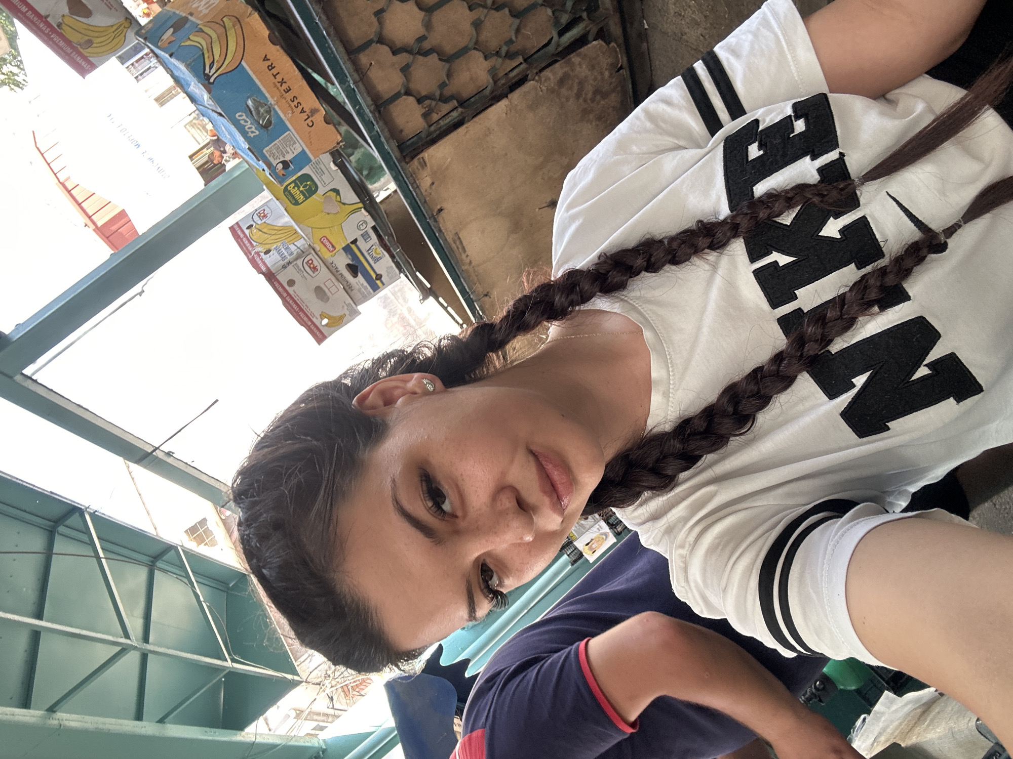 A young woman with dark hair in two braids is taking a selfie. She is wearing a white Nike t-shirt. Behind her, there are stacks of banana boxes. To her left, another person is partially visible, wearing a dark blue t-shirt with red trim. The environment appears to be an outdoor market, possibly under a covered structure with a green metal ceiling visible. The scene suggests a casual setting in a city, likely Sliven, Bulgaria, given the user's prompt.