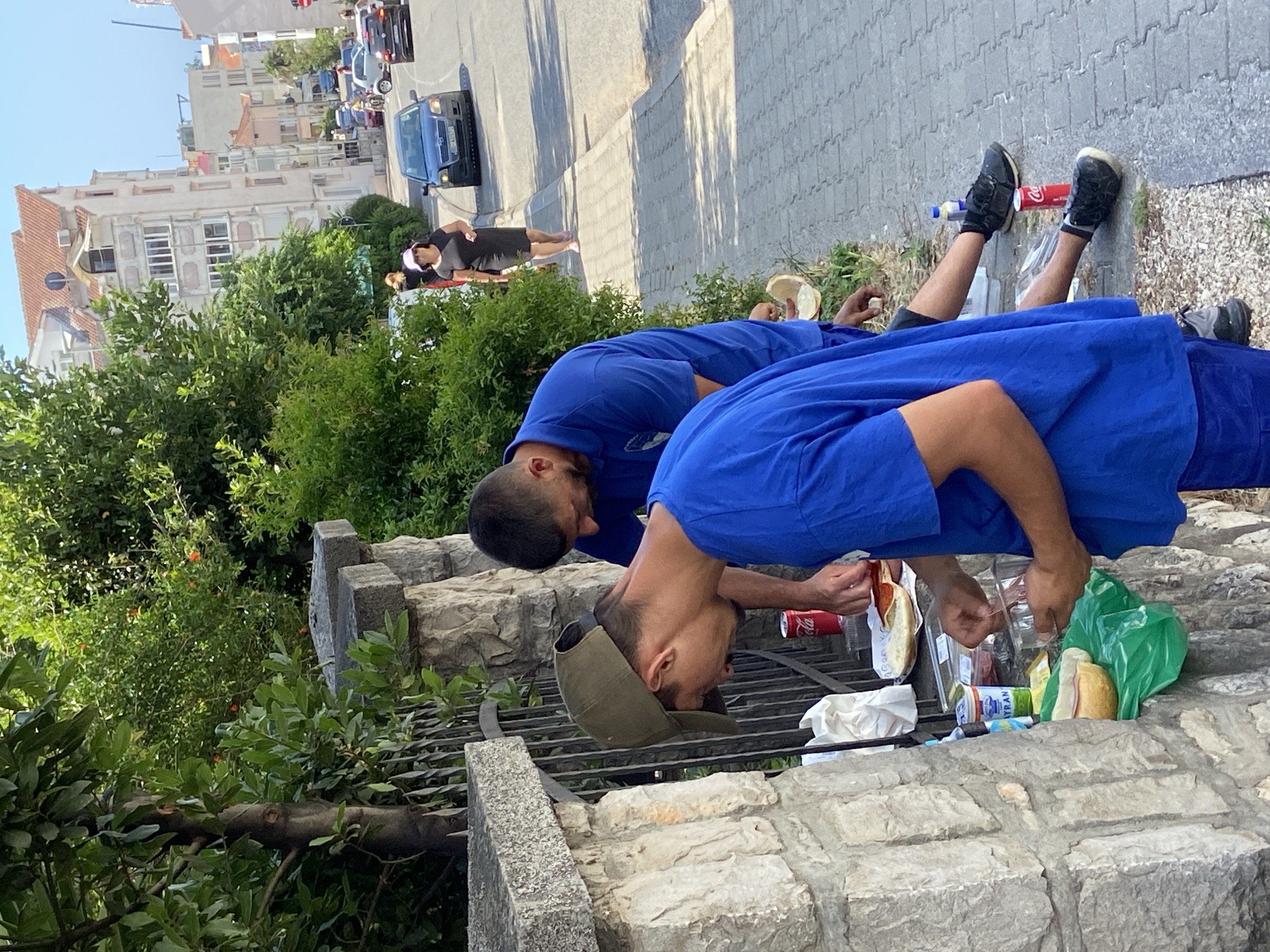 The image captures a street scene in Budva, Montenegro, on a sunny day. In the foreground, two men wearing blue shirts are preparing food outdoors next to a stone wall. The man on the left, wearing a green baseball cap, is intently focused on food items arranged on plates and in containers on the wall, which appears to be a makeshift preparation surface. He is holding a utensil and appears to be assembling food. To his right, another man in a blue shirt is also engaged in food preparation, reaching towards the items on the wall. Visible food items include bread, slices of meat, and possibly cheese. A red Coca-Cola can is placed near the food.
The environment includes a paved sidewalk to the right of the wall, with a street beyond it where vehicles are visible, including a black sedan and a white car. A woman in a black dress and a pink hat is walking on the sidewalk in the middle ground. Lush green trees and bushes line the street and the area behind the stone wall. In the background, apartment buildings with reddish-tiled roofs are visible, characteristic of urban architecture. The overall scene suggests a casual outdoor meal or break being taken by the two men amidst the urban environment of Budva.