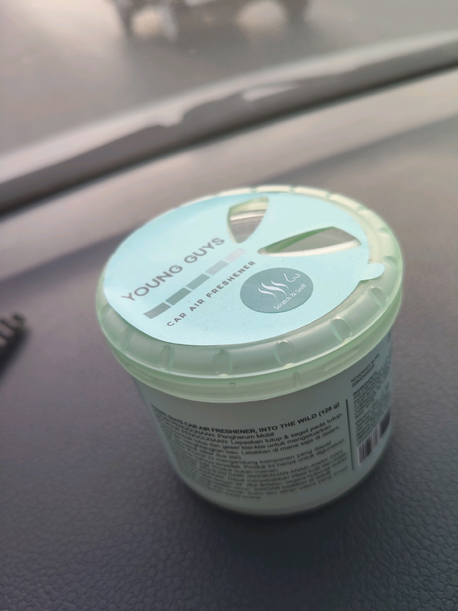 A close-up, angled shot shows a light blue car air freshener with the brand "YOUNG GUYS" printed on its lid in white text. Below the brand name, "CAR AIR FRESHENER" is also printed. To the right of this text, there is a small circular emblem with a wavy line design and the words "Scratch & Sniff" underneath. The lid has a perforated section that has been partially opened. The product is cylindrical and appears to be made of plastic, with a textured light green base. The product is resting on a dark, textured surface. The background is out of focus, showing indistinct gray and white shapes that suggest an indoor setting, possibly a car interior due to the product's nature. There are no people visible in the image. The product's label contains text in Indonesian, including "PENGHARUM MOBIL" (Car Freshener) and instructions for use. The scene is lit by ambient light, casting soft shadows.