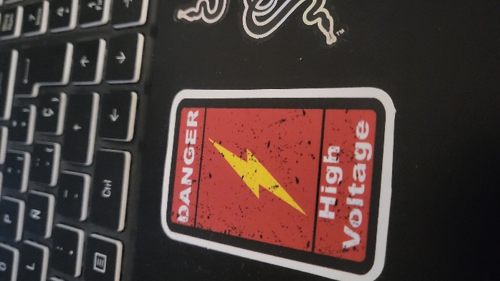 The image displays a close-up, angled view of a section of a dark-colored laptop. A rectangular sticker, featuring a red background with black distressed textures and a white border, is affixed to the laptop's palm rest area. The sticker prominently displays the word "DANGER" in white uppercase letters at the top, a yellow lightning bolt symbol in the center, and the phrase "High Voltage" in white uppercase letters at the bottom. Above the sticker, a portion of the laptop's black keyboard is visible, showing keys with white legends, including "Ctrl" and various arrow keys. To the right of the red sticker, a partial white outline of a second sticker, consistent with the Razer brand logo, is present on the laptop surface. No people are depicted. The scene occurs indoors under ambient lighting. The image provides no visual information to determine its specific location within the city of Magaluf, Spain, or any other geographical area.