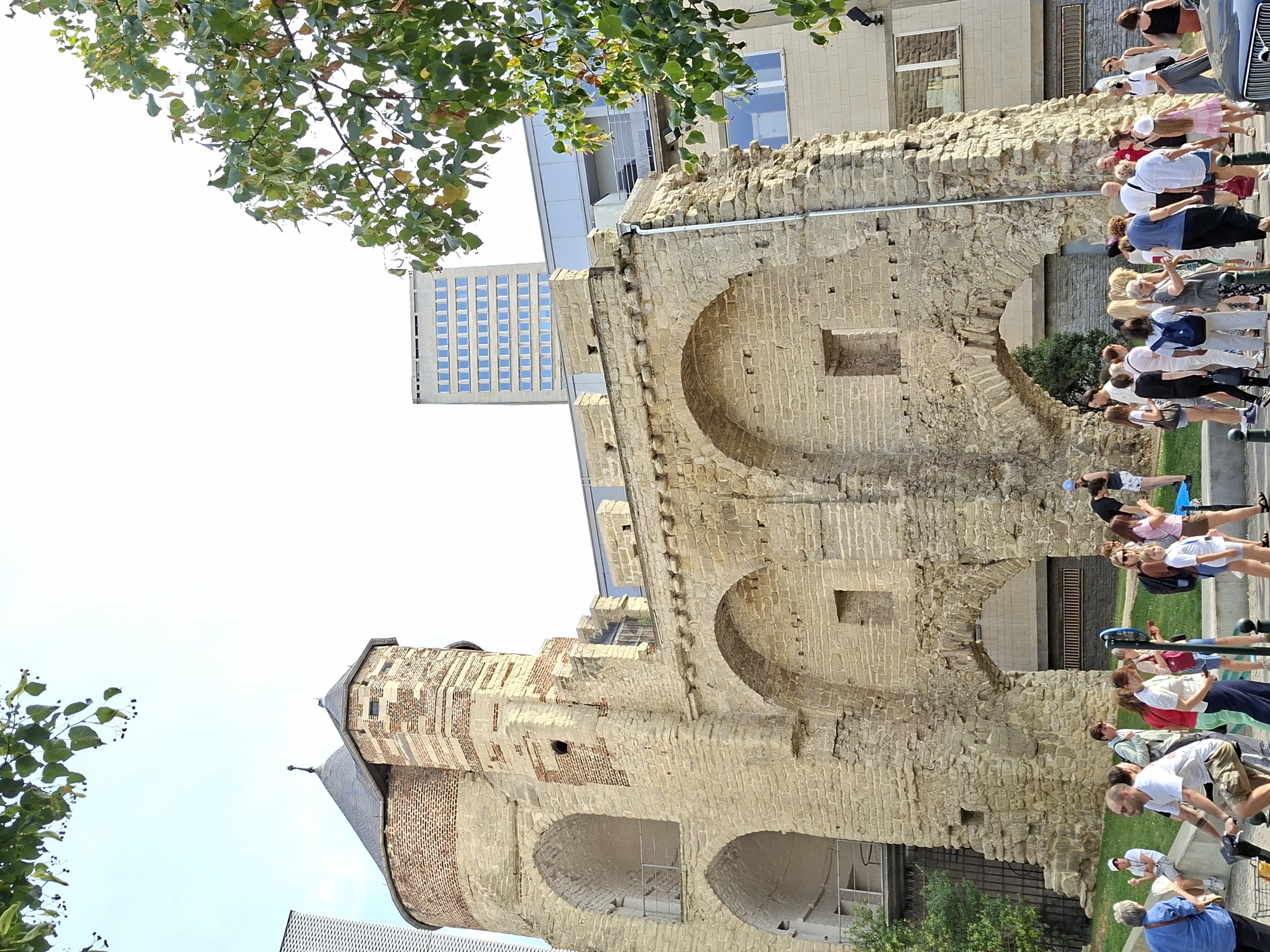 The image shows a street scene in Brussels, Belgium, featuring a historic stone gatehouse and tower, likely a remnant of the city's medieval fortifications. The ancient structure, made of light-colored stone blocks, exhibits arched openings and weathered brickwork, with one tower capped by a dark, conical roof. In the background, modern buildings, including a tall, rectangular structure with numerous windows, contrast with the historical architecture. A group of people, dressed in casual summer clothing, are gathered around the gatehouse and on a paved walkway. Some are standing and talking, while others are walking or looking at their phones. One person is riding a scooter. Green trees with some foliage are visible, framing the scene and adding to the urban park-like atmosphere. The sky is bright and appears to be clear.