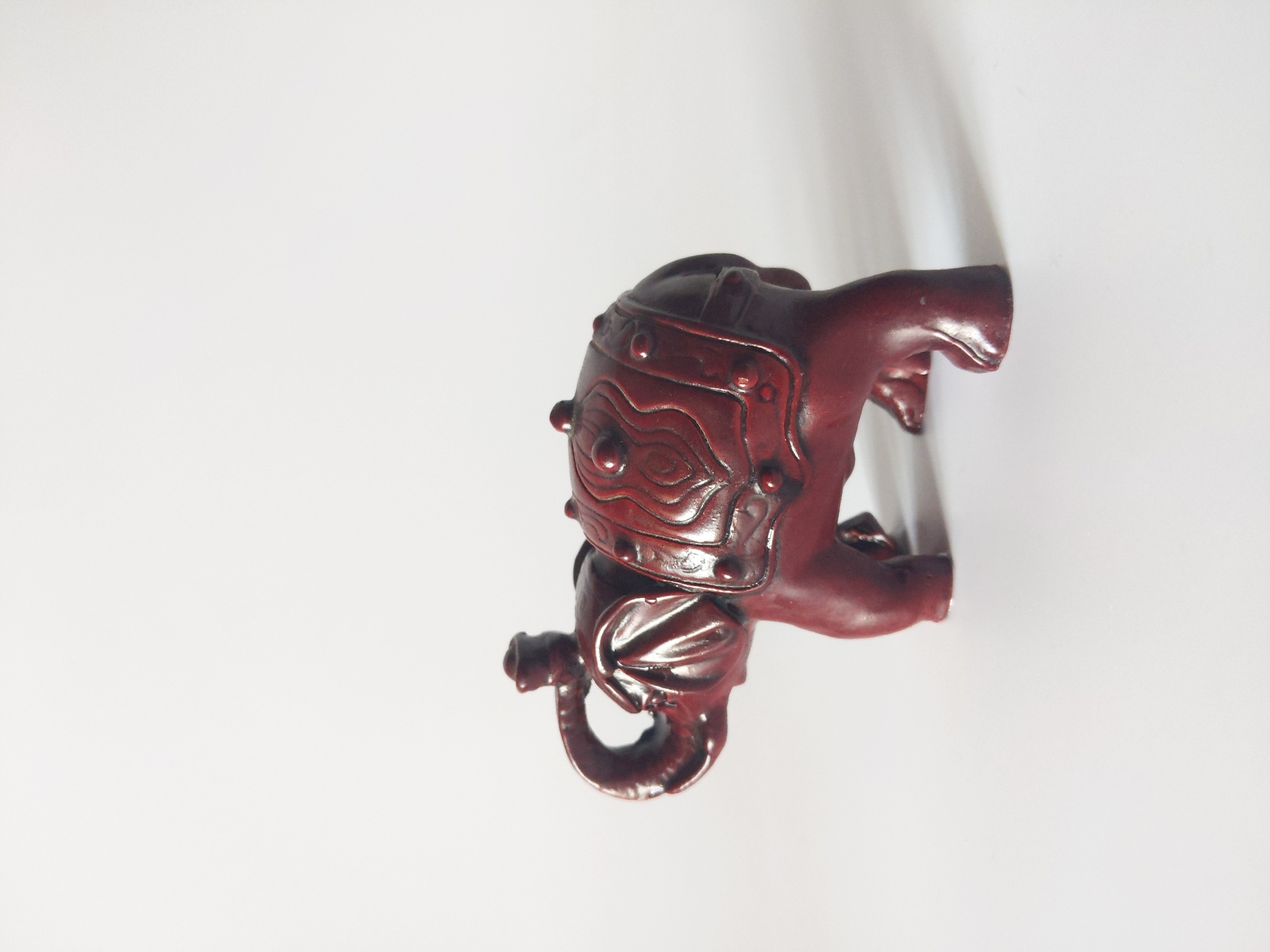 A dark red, carved wooden elephant figurine is placed on a plain white surface. The elephant is facing left, with its trunk curled upwards. It has decorative carvings on its back, resembling a saddle or blanket, with raised circular elements. The figurine has four legs, with the rear right leg slightly forward of the others. The lighting suggests it is indoors, with a shadow cast to the right of the elephant. The background is a solid white. The image's location in Rivne, Ukraine, is not discernible from the visual elements present in the photograph.