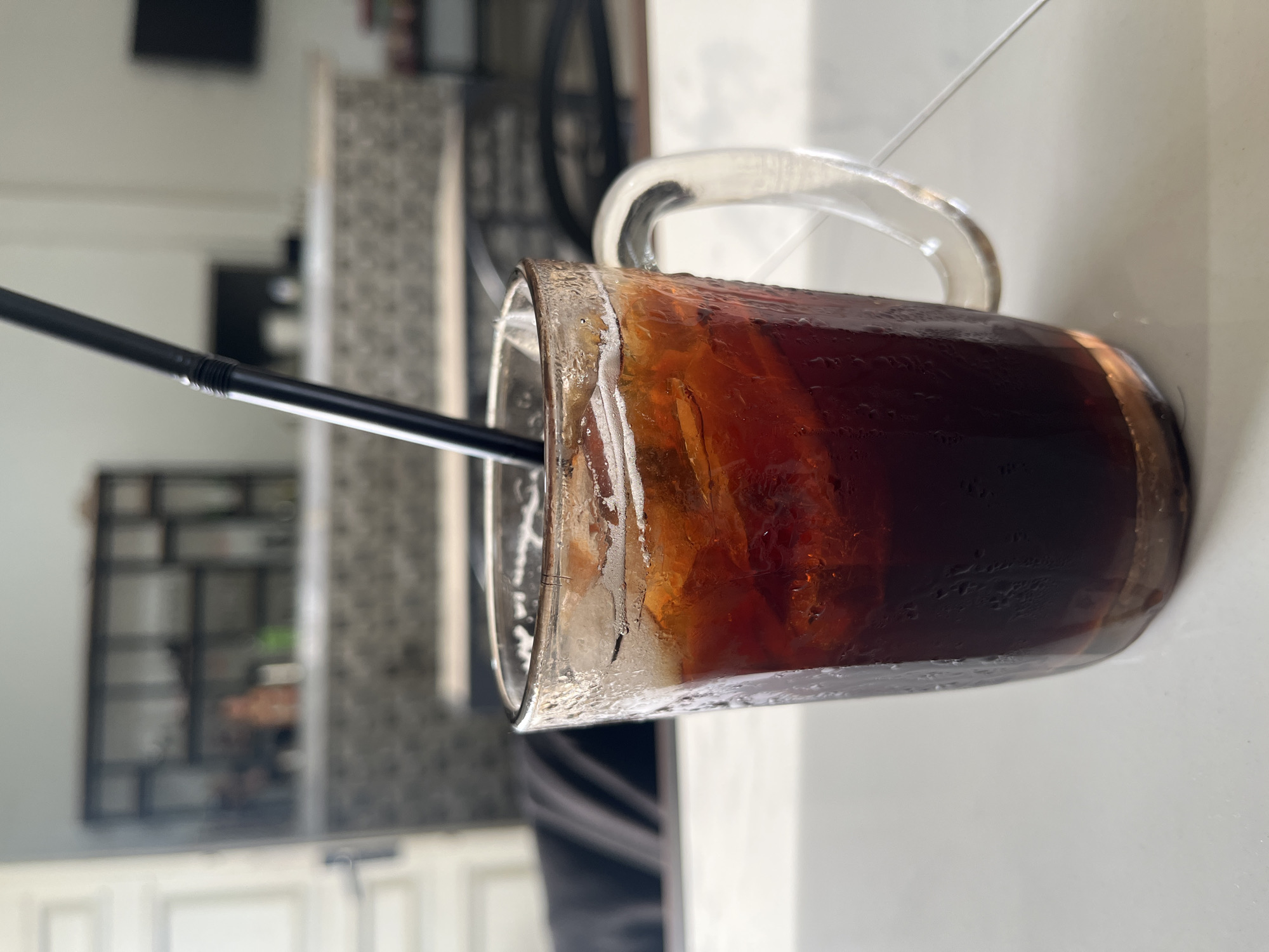 A close-up shot focuses on a glass mug filled with iced coffee and a black straw. The mug is on a white tiled surface, likely a table, in what appears to be an indoor seating area, possibly a cafe or restaurant. The background is blurred, showing shelves with various items, a counter, and a television mounted on the wall. The lighting suggests it is daytime. The location is identified as the city of Bireuenmeunasahreuleut, Indonesia.