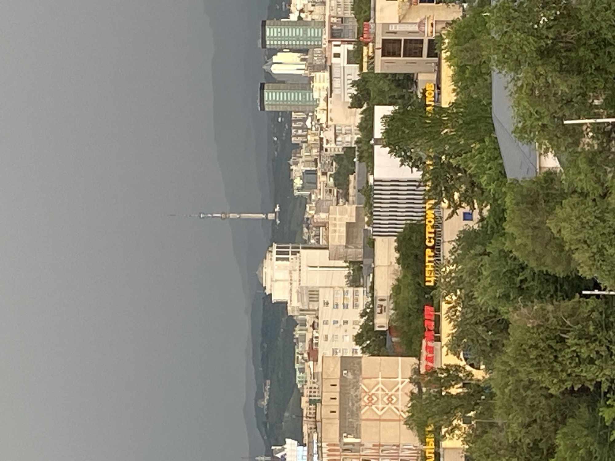 This media file depicts a city skyline with a prominent television tower. The scene is dominated by a dense array of buildings, varying in height and architectural style, suggesting an urban environment. Lush green trees are interspersed throughout the foreground and mid-ground, partially obscuring some of the structures and adding a natural element to the cityscape. The background features hazy, layered mountains, hinting at a mountainous region surrounding the city.
The image appears to be taken during the daytime, likely late afternoon or early evening, given the soft, diffused light and the muted colors. The sky is overcast, with a grayish hue, suggesting cloudy or possibly stormy weather.
Several signs with text are visible on the buildings. One sign in yellow letters reads "ЦЕНТР СТРОЙ". Another sign in red letters is partially visible and appears to say "АЗИАТ". These are Cyrillic inscriptions.
The overall impression is one of a bustling city nestled within a natural landscape, possibly during a period of inclement weather. The perspective is from an elevated viewpoint, looking down and across the urban sprawl towards the distant mountains. The image does not show any people or specific events taking place. The location is identified as Almaty, Kazakhstan.