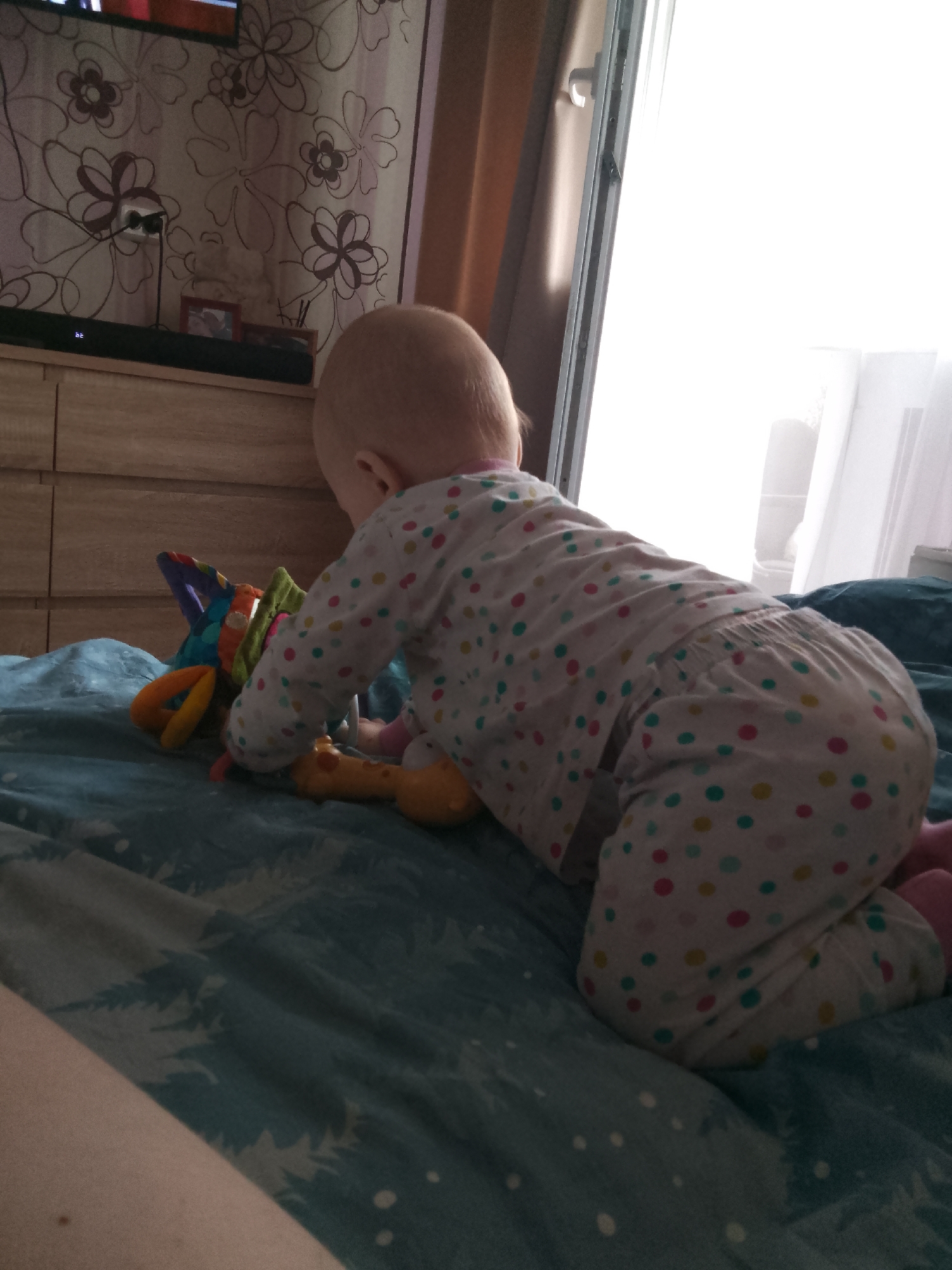 A baby is crawling on a bed with blue bedding, looking at a colorful toy. The baby is wearing pajamas with polka dots. The room has floral wallpaper and a dresser. A window with curtains is visible in the background. The image does not provide enough information to determine the location within the city of Minsk, Belarus.