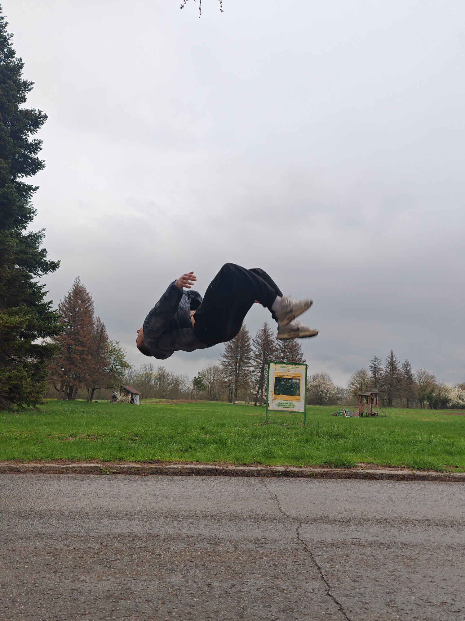 A person is in mid-air performing a flip, presumably a backflip. They are wearing a dark jacket, dark pants, and light-colored sneakers. The location is outdoors in a park area in Shumen, Bulgaria. The sky is overcast. Behind the person, there is green grass, scattered trees (including at least one evergreen), a sign with text (possibly a park information board), and playground equipment in the distance. In the foreground, there is a road with cracks in the asphalt.