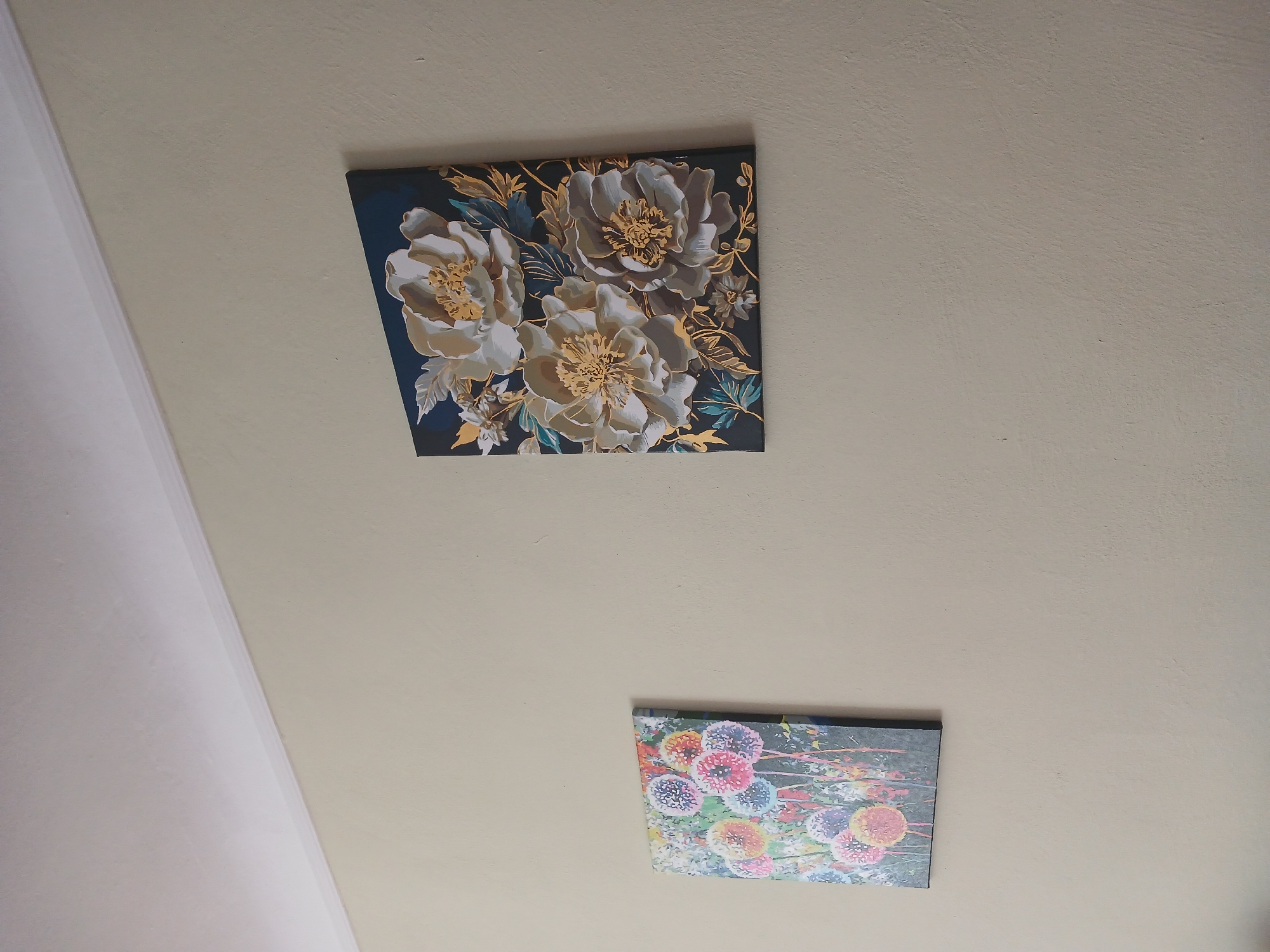 Two framed artworks hang on a light beige wall. The artwork on the left is a vertical canvas depicting colorful, abstract dandelion-like seed heads. The artwork on the right is a square canvas featuring a close-up of three large, stylized white and gold flowers against a dark blue background. The wall is capped by a white crown molding and a white ceiling is visible at the top of the frame. The image appears to be an interior shot, possibly a home or an office, located in Mamaivtsi, Ukraine.