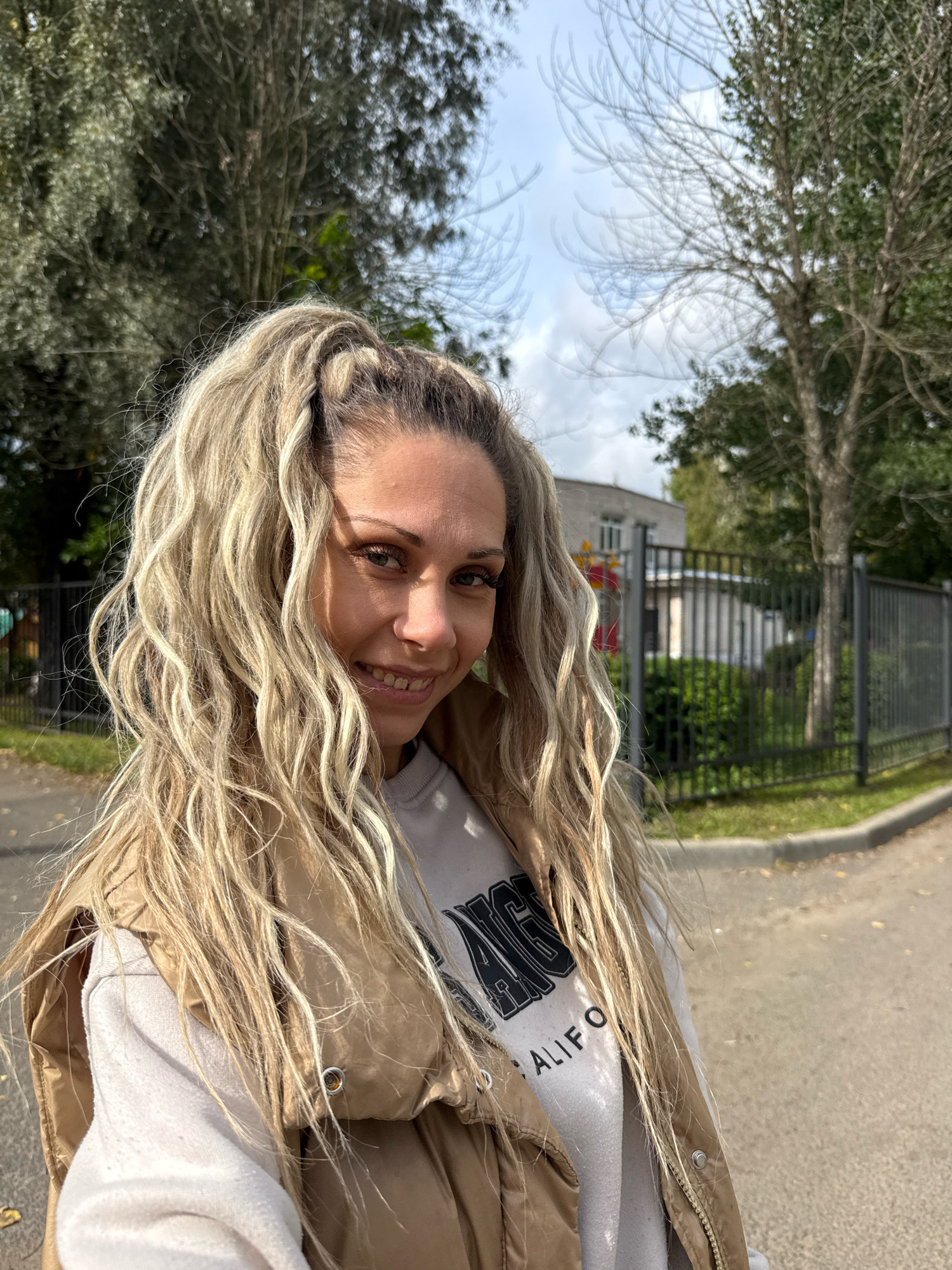 A woman with long, blonde, wavy hair pulled into a high ponytail smiles at the camera. She is wearing a tan puffer vest over a light-colored sweatshirt with "CALIFORNIA" printed on it. She appears to be standing outdoors on a paved path, with trees and a fence in the background. A building is visible behind the fence. The lighting suggests it is daytime. The location is identified as Rusanovo, Russia.