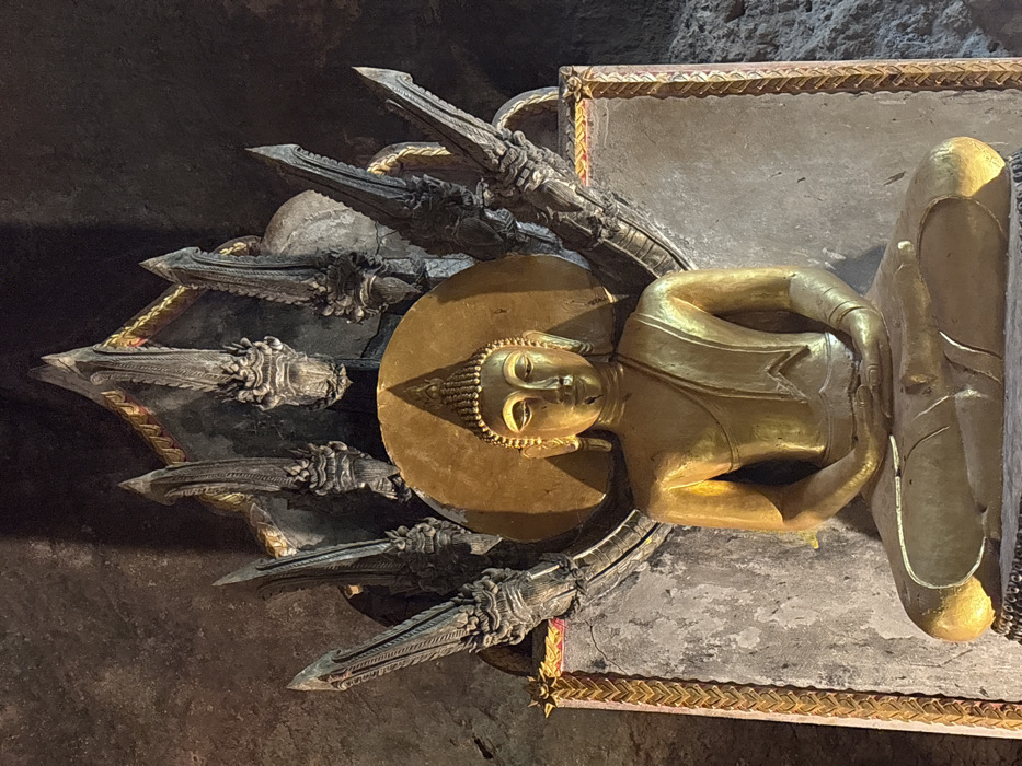 The image displays a golden Buddha statue centrally positioned within a dimly lit cave or rock temple, located in Ban Hin Sam Kon, Thailand. The Buddha is depicted in a seated meditative posture (Dhyana Mudra), with legs crossed and hands resting in the lap, palms upward. The statue has a radiant gold finish, featuring a serene expression, an ushnisha on the crown of the head, and elongated earlobes. A circular golden aureole with a pointed top frames the Buddha's head. Behind the Buddha, a large, dark grey stone or concrete relief forms a multi-headed Naga (cobra) canopy, with at least seven distinct cobra heads fanning out. This Naga structure exhibits detailed carvings of scales and heads, with subtle gold accents along its base and edges. The surrounding environment consists of uneven, dark rock walls, indicating a natural cave or a structure carved directly into rock. The Buddha statue is illuminated by direct light, contrasting with the darker background.