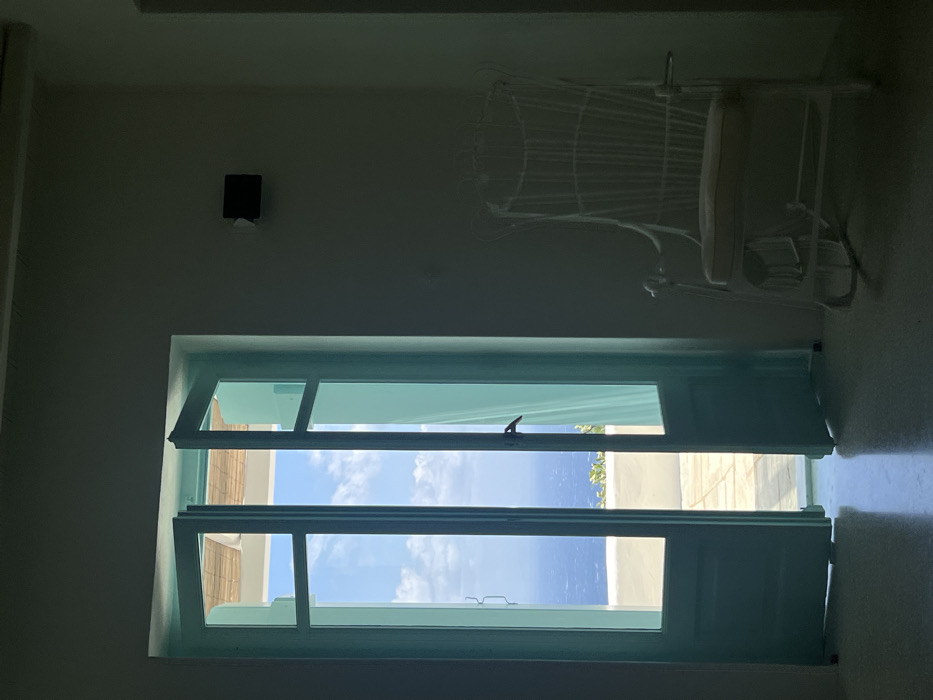 This image depicts an interior room in Tourlos, Greece, looking out through an open set of turquoise double doors to a bright exterior. The doors, featuring multiple glass panes, reveal a light-colored patio with a low wall and green foliage, beyond which lies a wide expanse of deep blue sea under a sky with scattered clouds. A segment of a light-colored, possibly thatched, outdoor canopy structure is visible at the top of the exterior view. Inside the predominantly shadowed room, a white, high-backed, wicker-style rocking chair with a light-toned cushion is positioned to the right of the doorway. A small, dark, rectangular object is affixed to the wall above the chair. The reflective floor of the room shows a bright illumination from the exterior. No individuals are present in the scene, and no actions or interactions are depicted.