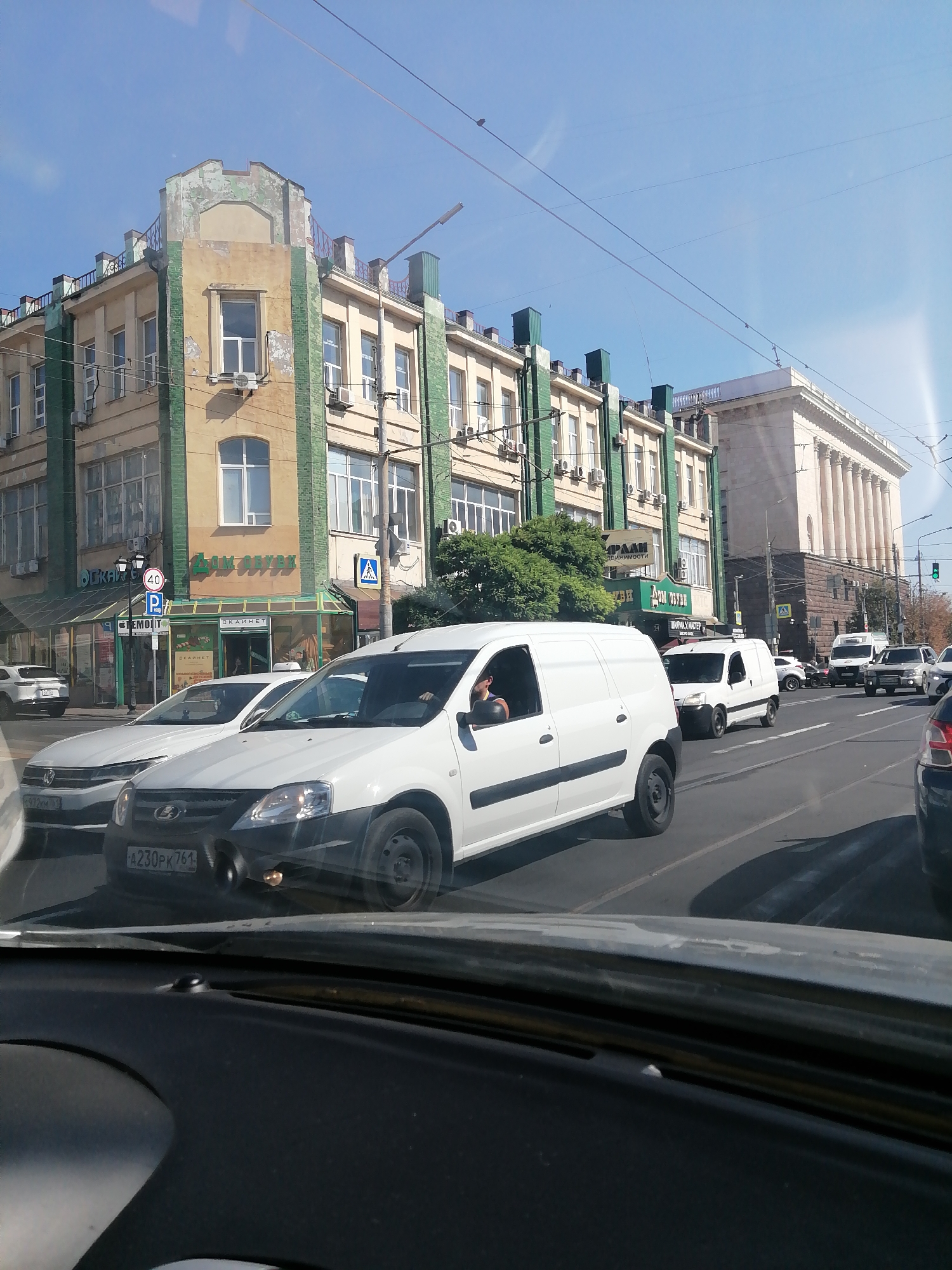 The image shows a street scene in Rostov-on-Don, Russia, on a sunny day. In the foreground, the dashboard of a vehicle is visible. Outside the vehicle, a white van with a driver is traveling on a multi-lane road. Several other vehicles, including cars and vans, are also present on the road. On the left side of the road, a light-yellow building with green trim and multiple windows stands. A sign on the building reads "Дом Обуви" (House of Shoes). Further down the street, a large building with classical architecture and columns is visible. Power lines crisscross the sky. The overall environment suggests an urban setting with moderate traffic.