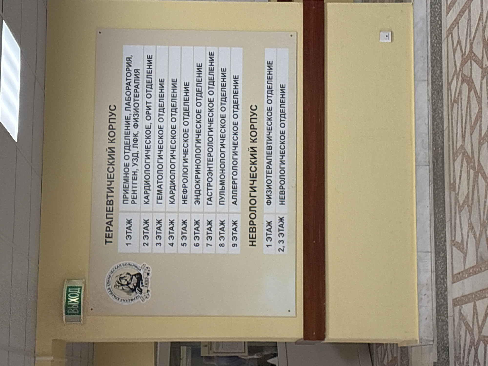 This is a photograph of a sign within a medical facility, detailing the floor-by-floor layout of the Therapeutic and Neurological buildings. The sign, mounted on a pale yellow wall, lists the departments and their respective floors within each building.
The sign for the Therapeutic building lists the following:
- Floor 1: Reception, Laboratory, X-ray, Ultrasound, Physical Therapy
- Floor 2: Cardiology, Resuscitation
- Floor 3: Hematology
- Floor 4: Cardiology
- Floor 5: Nephrology
- Floor 6: Endocrinology
- Floor 7: Gastroenterology
- Floor 8: Pulmonology
- Floor 9: Allergology
The sign for the Neurological building lists the following:
- Floor 1: Physiotherapy
- Floors 2, 3: Neurology
To the left of the sign, there is a lit-up "EXIT" sign and the emblem of Perm Regional Hospital with the year 1833. This indicates that the medical facility is located in the city of Perm, Russia.