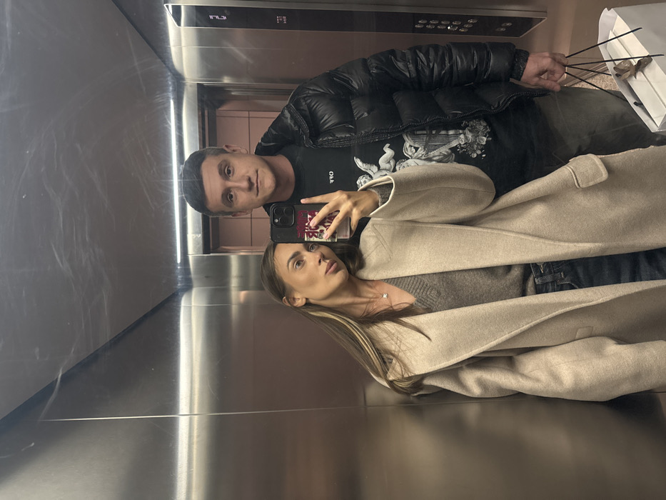 A couple is reflected in a mirrored elevator. The man wears a black puffer jacket and a black t-shirt. The woman, holding the phone taking the picture, has on a grey sweater and a tan coat. Both are angled towards the camera. A shopping bag and incense sticks are visible in the upper right corner. The elevator’s metal walls and buttons are evident in the background and foreground.
