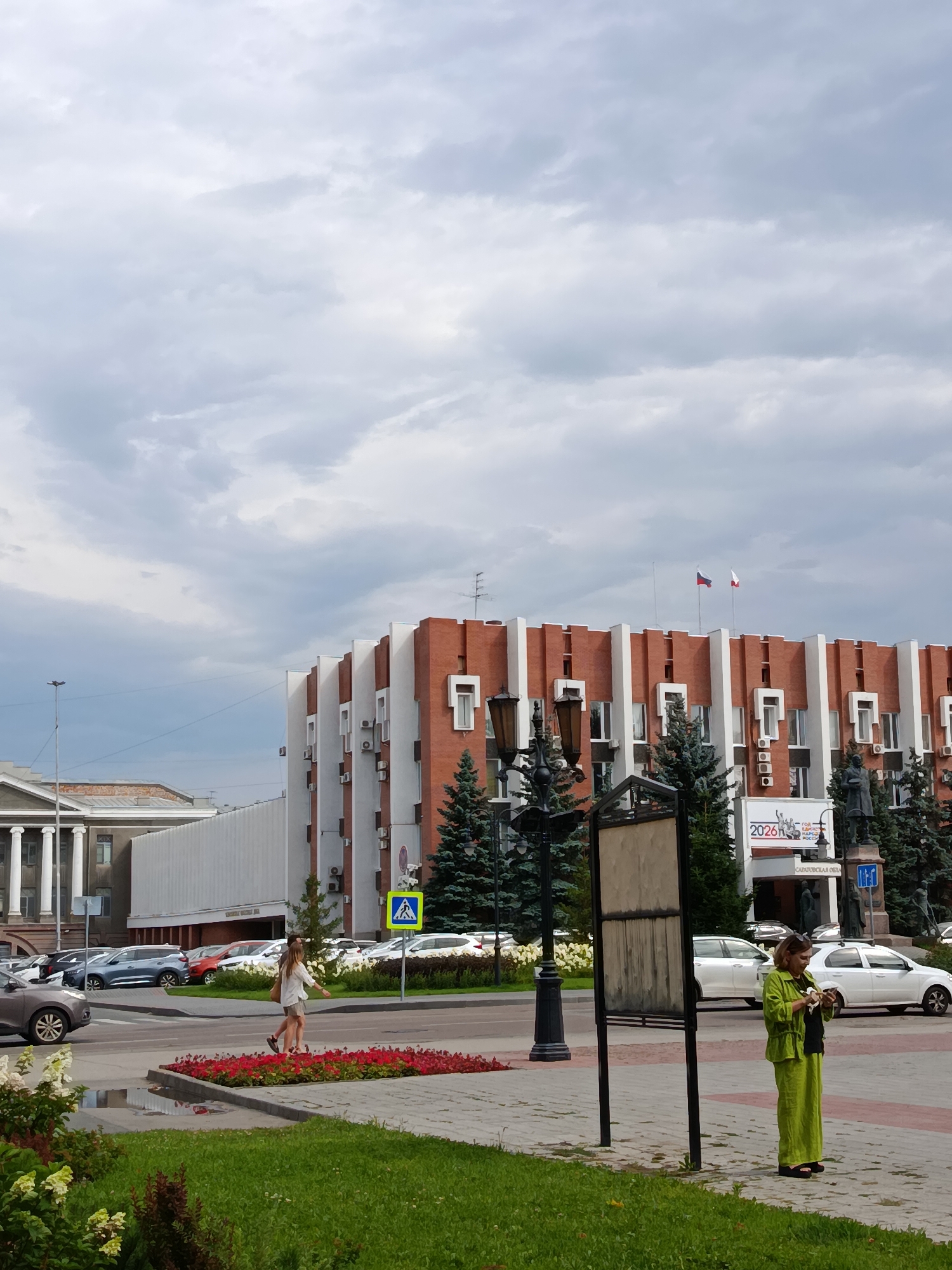 The image depicts a street scene in Saratov, Russia, with a focus on a building with a distinctive red brick and white facade. Two women are visible: one in a green outfit stands near a billboard and checks her phone, while another woman in lighter clothing walks across a pedestrian crossing. A row of cars is parked along the street, and a few trees are scattered throughout the scene. The sky is overcast with clouds. In the background, a building with classical columns is visible. A sign with "2026" and Russian text is mounted on the red and white building.