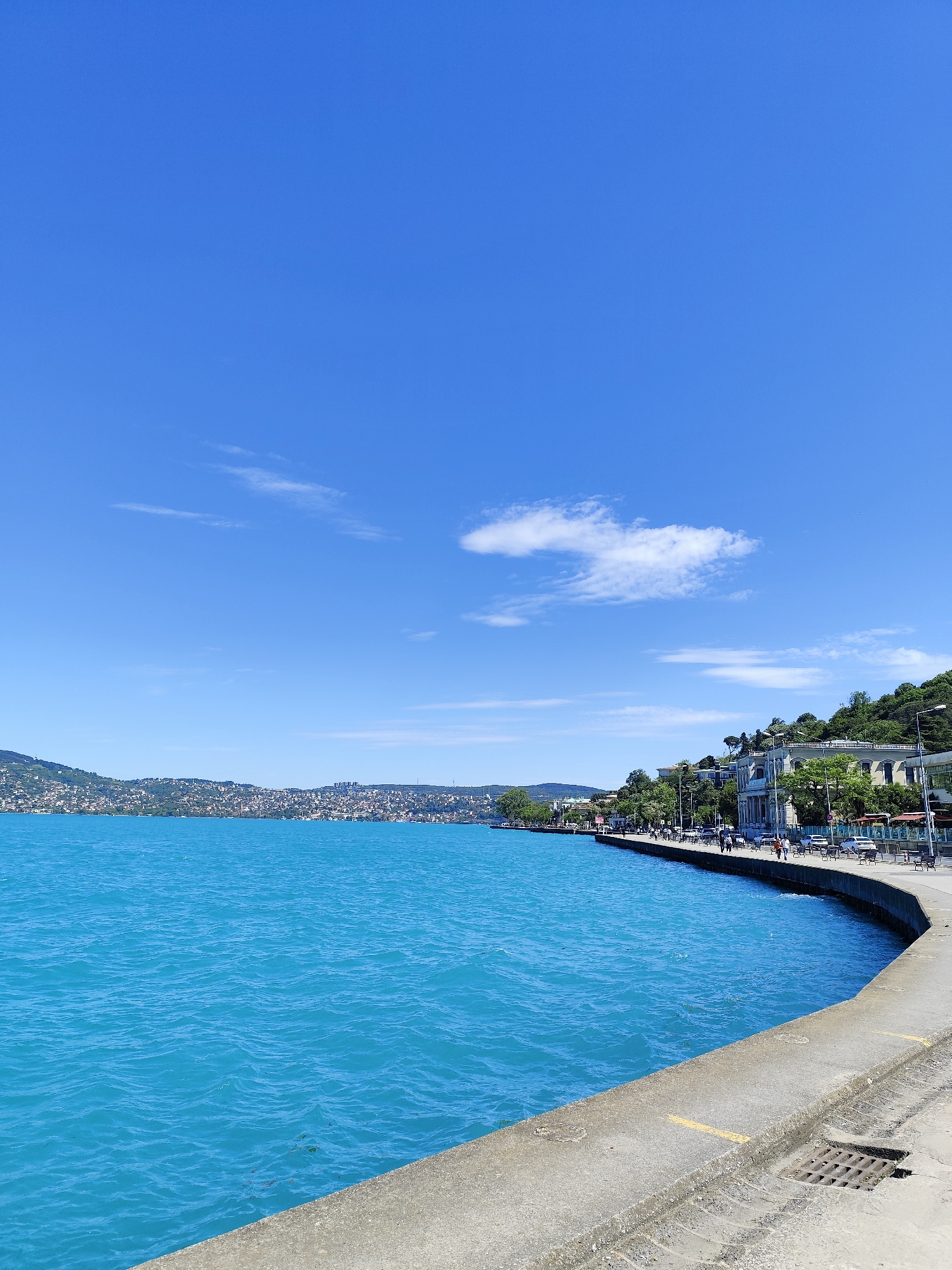 The image presents a view of the Bosphorus Strait from the shore in Sarıyer, Turkey. The sky is a clear, bright blue with scattered light clouds. The water is a vibrant turquoise blue, with slight ripples on the surface. Across the strait, the opposite shoreline is visible, densely populated with buildings climbing the hills.
Along the near shoreline, a concrete promenade curves along the water's edge. People are strolling and sitting on benches. Trees and vegetation line the promenade, and buildings are set back from the path. Streetlights and parked cars are visible. The foreground is dominated by the concrete edge of the promenade, showing signs of wear and containing a grated drain.