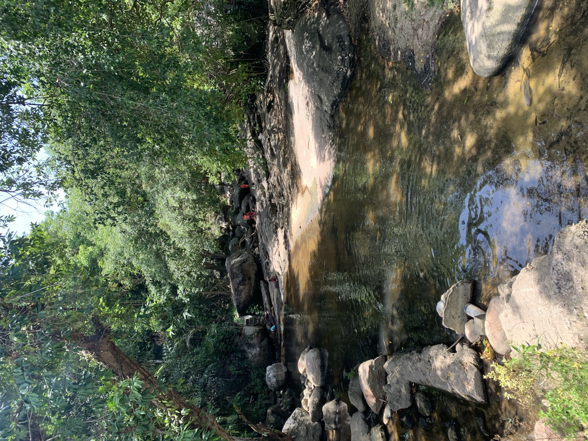 The image depicts a natural stream in a tropical environment, likely in the Ko Samui District of Thailand, characterized by lush green foliage and large rocks. The water is shallow and clear, revealing a sandy bottom in some areas. Several people are present, some wading in the stream and others sitting or standing on the rocky banks. Sunlight filters through the dense tree canopy, casting dappled shadows on the water and rocks. The scene suggests a recreational area, possibly a popular spot for locals and tourists to cool off and enjoy nature.