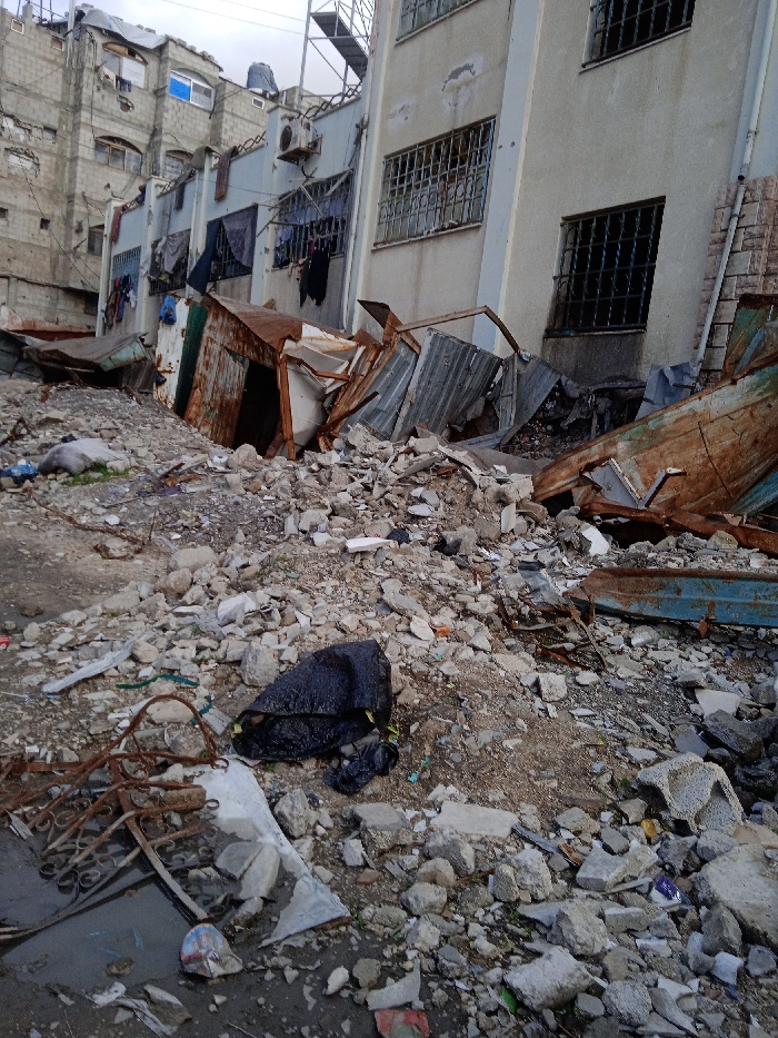 The image portrays an urban scene in Khan Yunis, Palestinian Territory, dominated by widespread destruction and debris under an overcast sky. The foreground and midground are covered by an extensive expanse of concrete rubble, broken masonry, dirt, and fragmented materials. Visible objects include a black plastic bag resting on the debris in the lower-center, and various pieces of rusted, twisted metal, including what appear to be collapsed structures or shelters made of corrugated iron sheeting, partially submerged within the rubble. In the background, two multi-story residential buildings stand amidst the damage. The building on the left is constructed from grey concrete blocks, exhibiting visible wear and damage, with multiple windows, some appearing boarded or covered, and clothes hanging from exterior lines. An air conditioning unit is mounted on its facade, and a metal structure is visible on the roof. The adjacent building on the right is a lighter beige/grey, featuring numerous windows secured with dark metal grates, and also displays some exterior damage to its plaster. Laundry hangs from the exterior of this building on its left side. No individuals are present in the depicted scene.