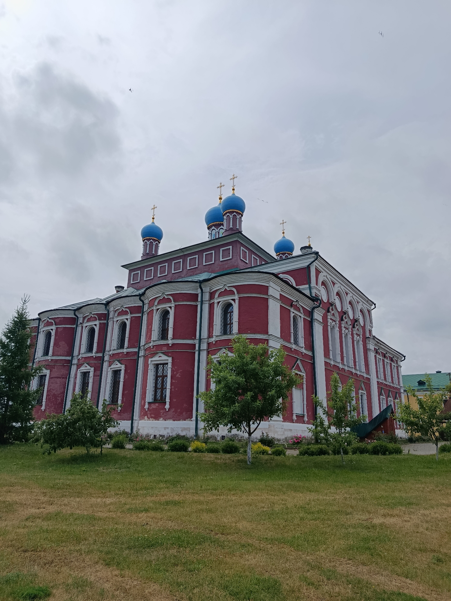 A large, red brick church with white trim and blue domes is situated in a grassy area with trees. The church has multiple windows, arched doorways, and a prominent cross on top of each dome. The sky is overcast with some clouds. In the distance, other buildings and trees are visible. The overall scene depicts an architectural structure in a rural or semi-rural setting under a cloudy sky.