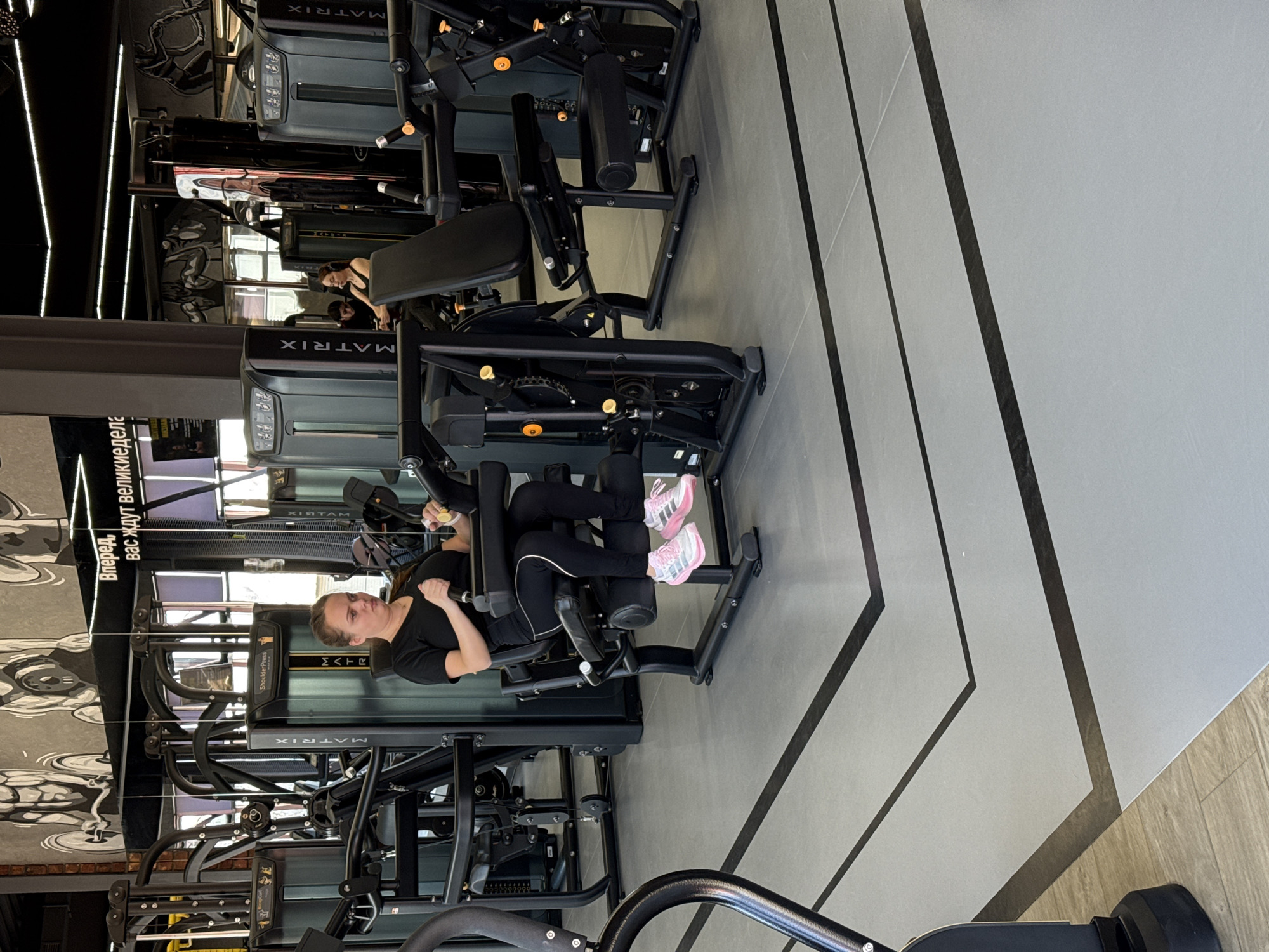 The image shows the interior of a gym, likely in Votkinsk, Russia, given the location information. A woman is using a seated leg curl machine with the "Matrix" brand visible on the equipment. She is wearing black athletic clothing and pink and white sneakers. The floor is gray with black geometric patterns. Other gym equipment is visible in the background, along with a text sign in Russian on a wall. The scene shows a person actively engaged in exercise using gym equipment.
