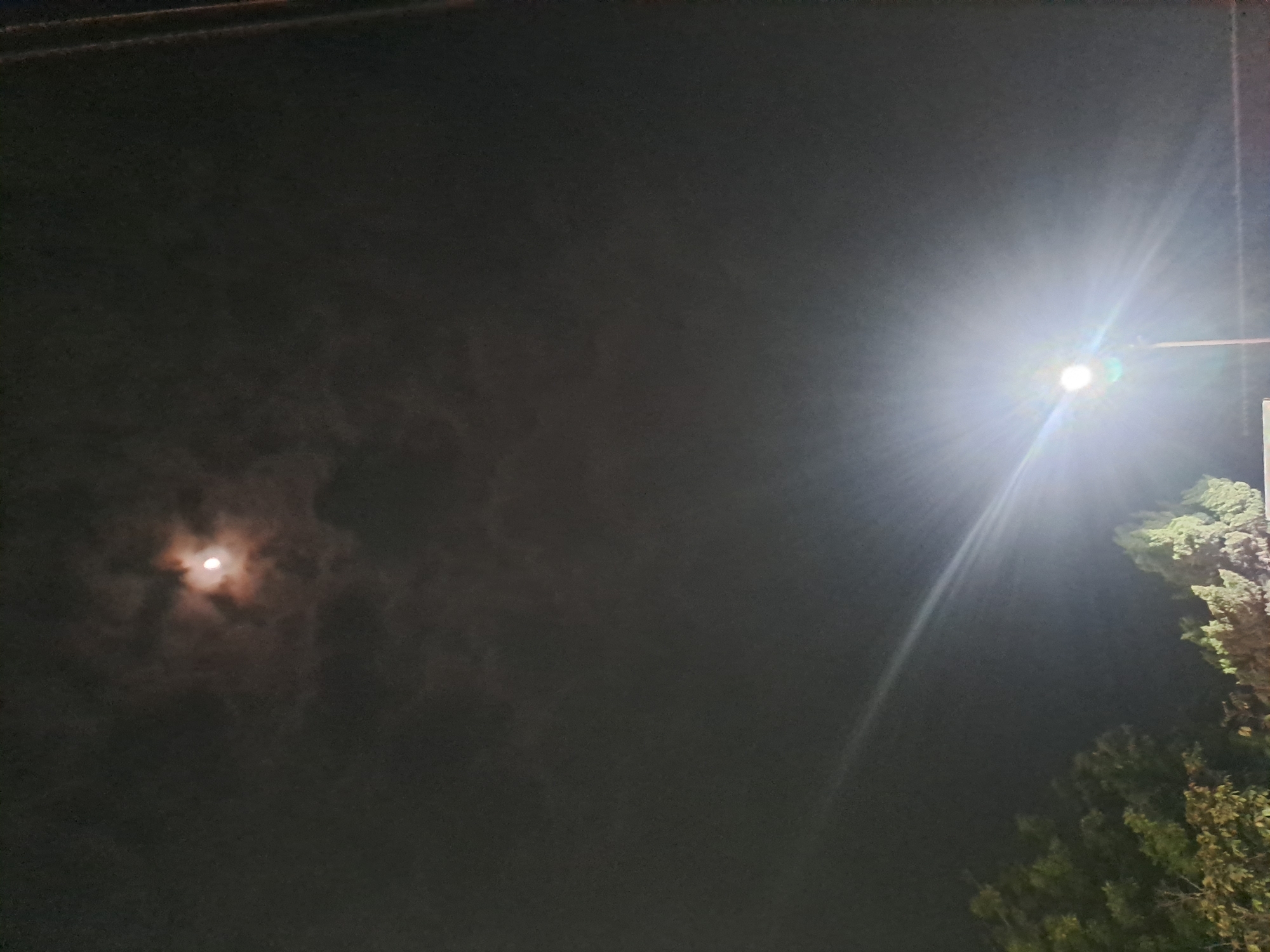 The image depicts a night sky with a bright moon partially obscured by clouds. In the lower right quadrant, a powerful street lamp illuminates the scene with a strong, white light, casting rays that streak across the sky. Below the lamp, the silhouettes of trees and possibly some low structures are visible. The overall impression is of a nighttime urban or suburban landscape, possibly in Slambur, Indonesia, where artificial light from the street lamp competes with the natural light of the moon. There are no people visible in the image.
