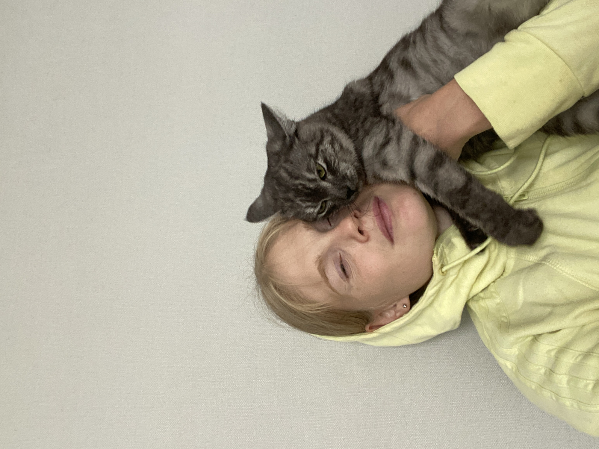 A close-up, waist-up shot shows a blonde woman wearing a pale yellow hooded sweatshirt, with a grey tabby cat being held against her face. The woman's eyes are mostly closed, and she is looking slightly upward. The cat's face is pressed against the woman's cheek, with its green eyes open and looking towards the viewer. The background is a plain, light-colored wall. The setting appears to be indoors. The image is a selfie, as indicated by the perspective and the framing. There is no information in the image to determine the location in Chaikovsky, Russia.