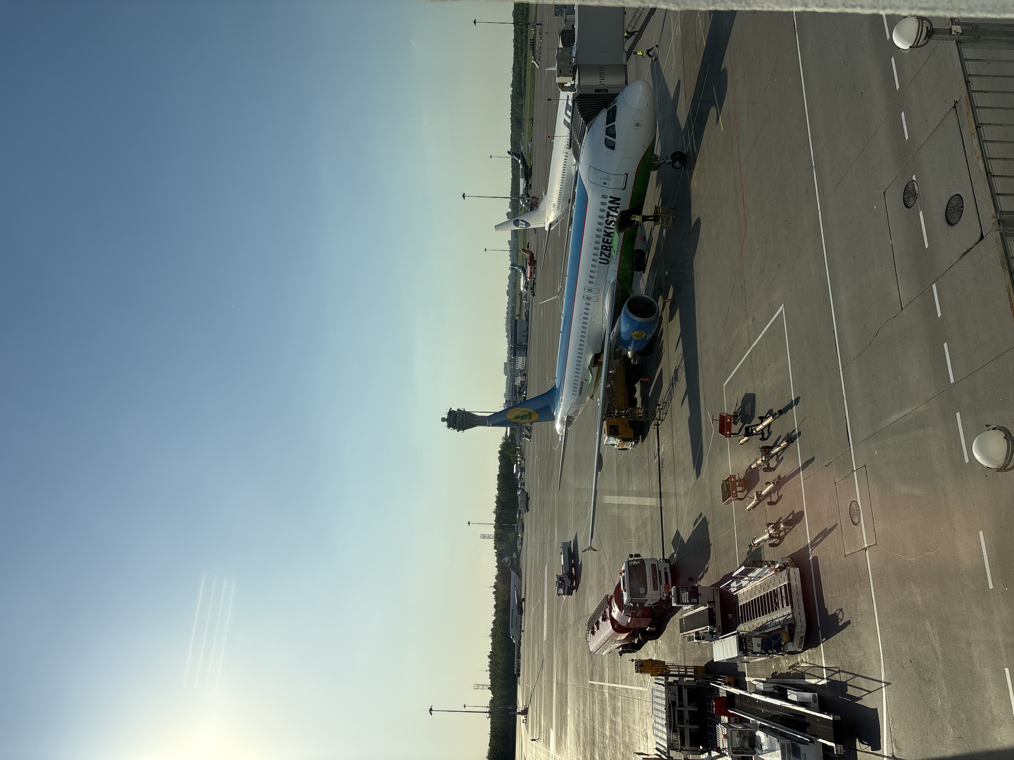 The image shows an aerial view of Pulkovo Airport in Saint Petersburg, Russia. There are two Uzbekistan Airways planes parked at gates, attended by ground crew. A fire engine and other service vehicles are parked nearby on the tarmac. The airport terminal and control tower are visible in the background. The sky is clear.