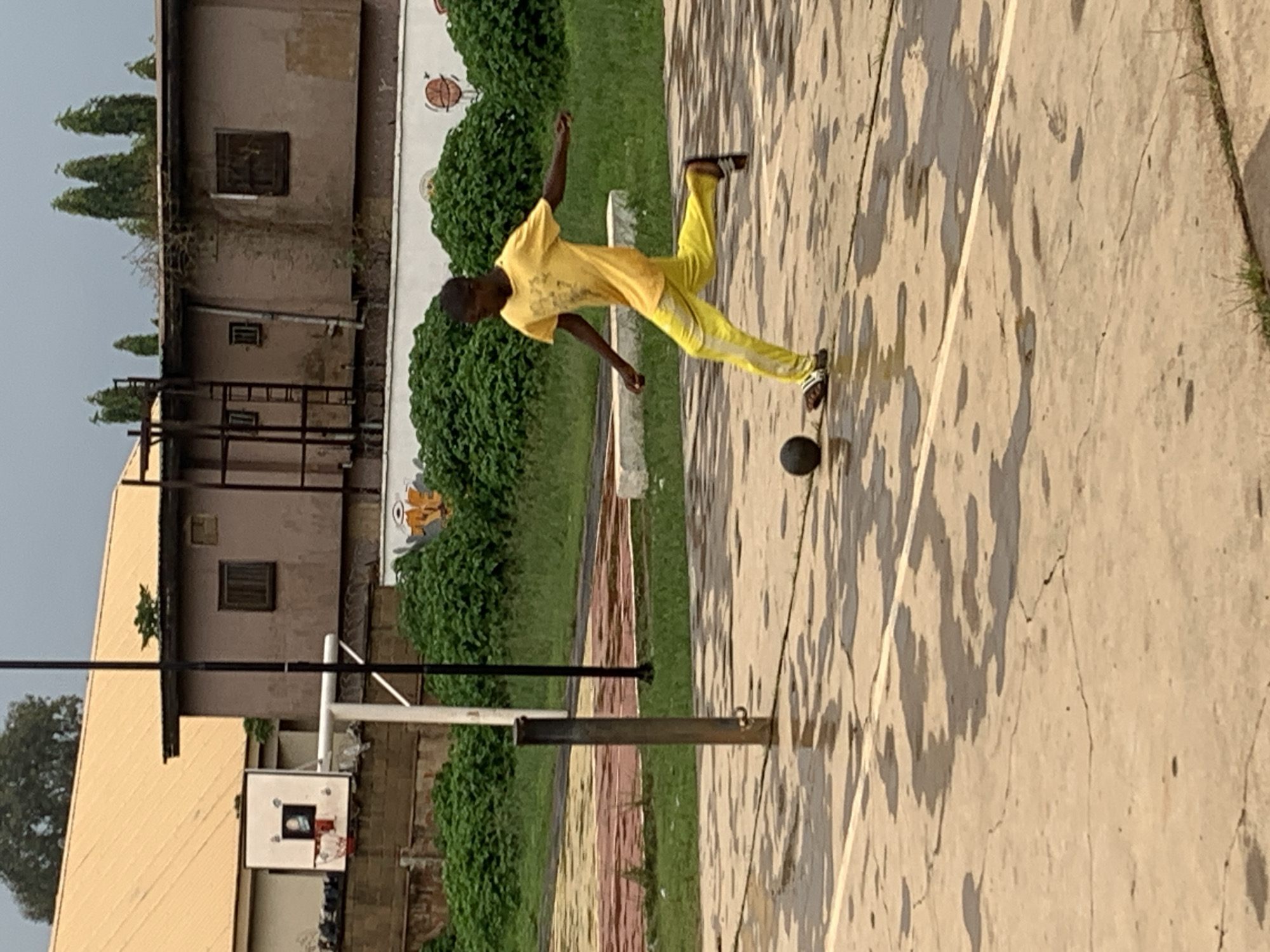 A young boy, appearing to be of African descent, is captured mid-action on a wet outdoor court, likely a basketball court based on the presence of a backboard and hoop in the background. He is wearing a bright yellow t-shirt with a graphic design and matching yellow pants with a white stripe down the side. The boy is in the process of kicking a dark-colored ball, his right leg extended forward. His left leg is bent, supporting his weight. He is wearing sandals.
The court surface is concrete and appears wet, with numerous puddles reflecting the overcast sky. A white line marks the boundary of the court. In the background, behind the court, is a thick, dense hedge of green bushes. Beyond the bushes, there are buildings. One building has a distinctive orange-tiled roof and a brown facade with several windows, one of which is barred. Another building to the right is also brown with a weathered appearance and a dark, boarded-up window. A metal ladder structure is visible attached to one of the buildings. A tall, thin metal pole stands prominently in the foreground, and a black pole is also visible to its right. The overall environment suggests an urban or semi-urban setting, possibly within the city of Jos, Nigeria, given the architectural style and the presence of tropical vegetation. The sky is a uniform grey, indicating cloudy or rainy weather.