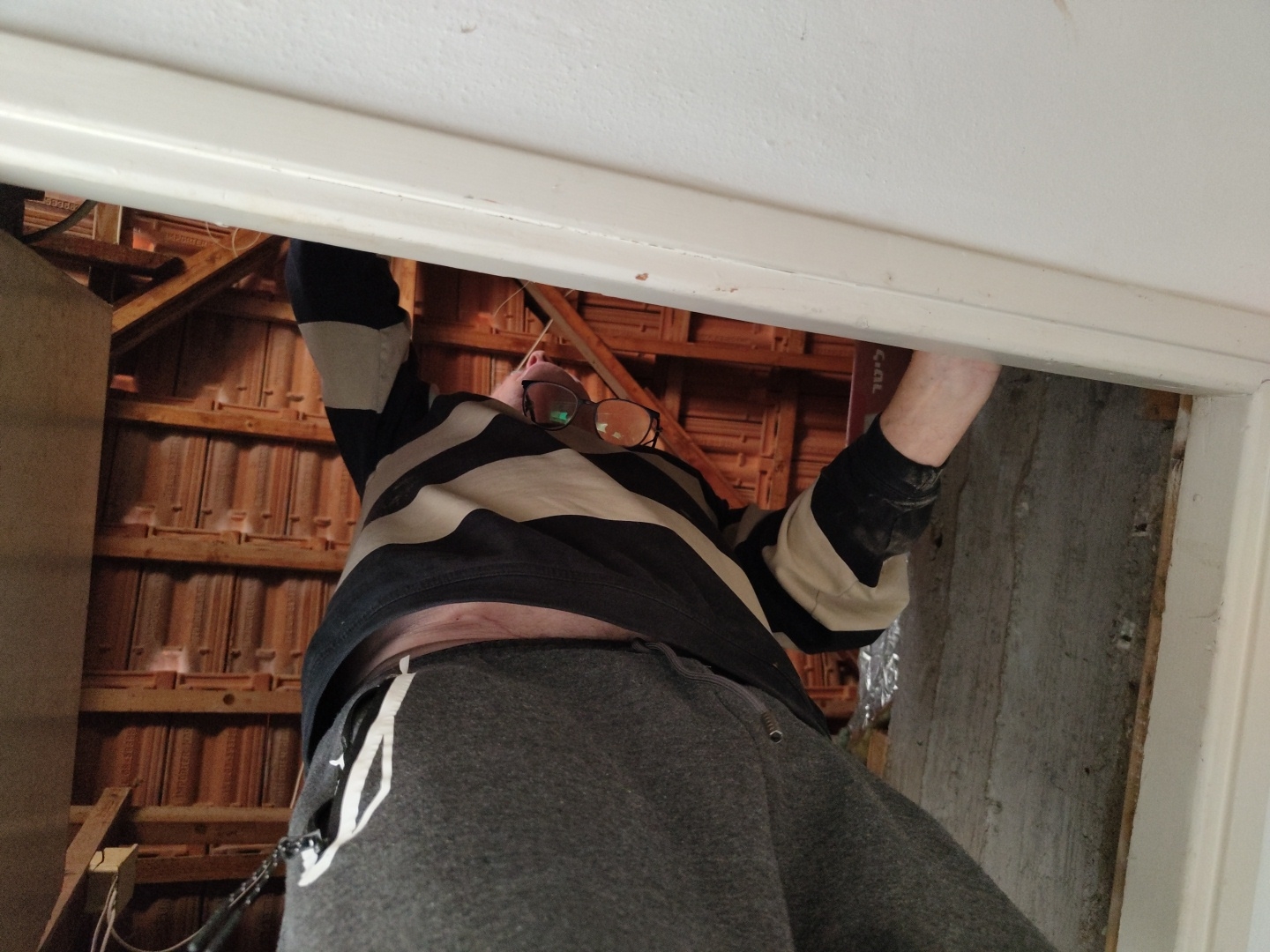 The image captures a partial view of an individual accessing an attic or crawl space in Raanana, Israel, from a low-angle perspective. The subject, identifiable from the waist up, is bent forward, entering the space. They are wearing grey sweatpants, a horizontally striped long-sleeved shirt with black and off-white/beige bands, and a dark sleeve or glove on their left arm. A pair of eyeglasses with dark frames and orange-tinted lenses hangs from the shirt's collar. A metallic chain is visible attached to the left side of their sweatpants. The person's bare right forearm and hand are extended into the space, holding a red object. Partially visible Hebrew text on this object reads "ליטר" (liter). The environment beyond the white-framed opening consists of an unfinished attic space with exposed wooden beams and reddish-brown undersides of roof tiles in the background. The floor of the attic appears to be a rough, light grey concrete-like surface, with some silver foil insulation visible.