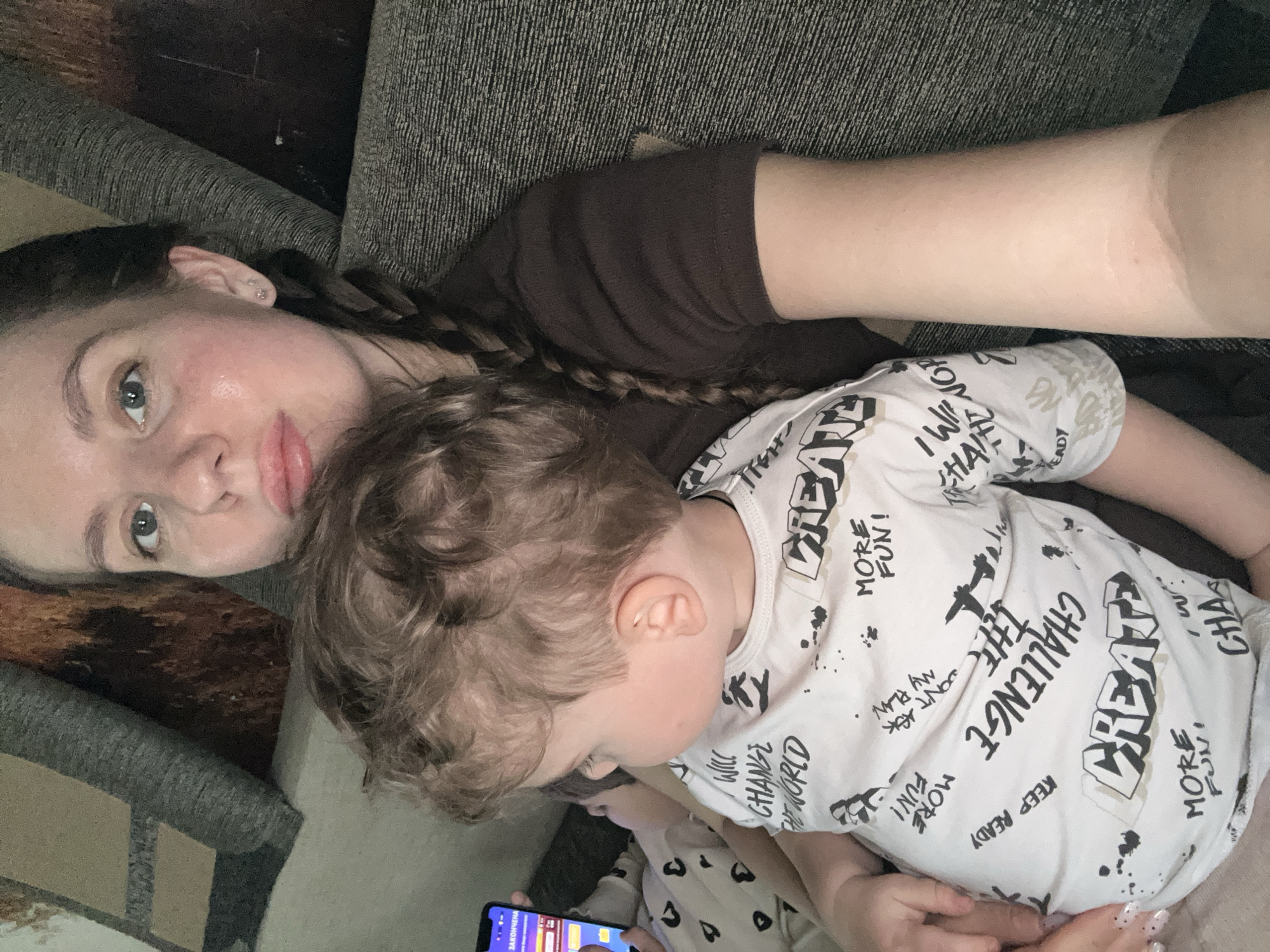A woman with brown hair braided to the side is lying on a gray couch next to a young child with curly hair. The woman is looking at the camera. The child is holding a phone. The child is wearing a white t-shirt with black graphic text. The woman is wearing a brown t-shirt. The location is not discernible from the image.