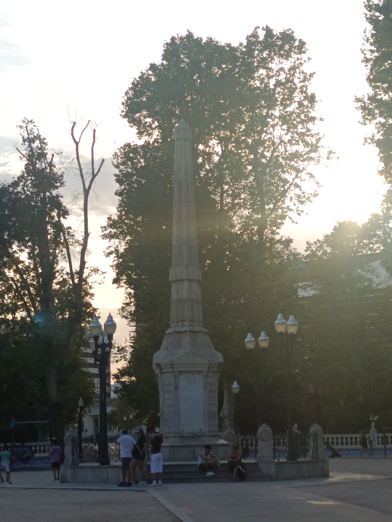 A tall, ornate monument, possibly a memorial or obelisk, stands at the center of the scene, bathed in the warm, hazy light of a sunset. The monument is surrounded by trees that are silhouetted against the bright sky, with sunbeams filtering through their branches. Several lampposts are visible, adding to the public space atmosphere. A group of people are scattered around the monument, some standing and talking, others sitting on the monument's base or nearby steps. There are also children playing in the foreground, suggesting a park or public square setting. The architecture in the background appears to be traditional European style, with buildings and possibly a fence or railing. The scene is likely located in a public area of Castelló de la Plana, Spain, given the context of the monument and surrounding environment.