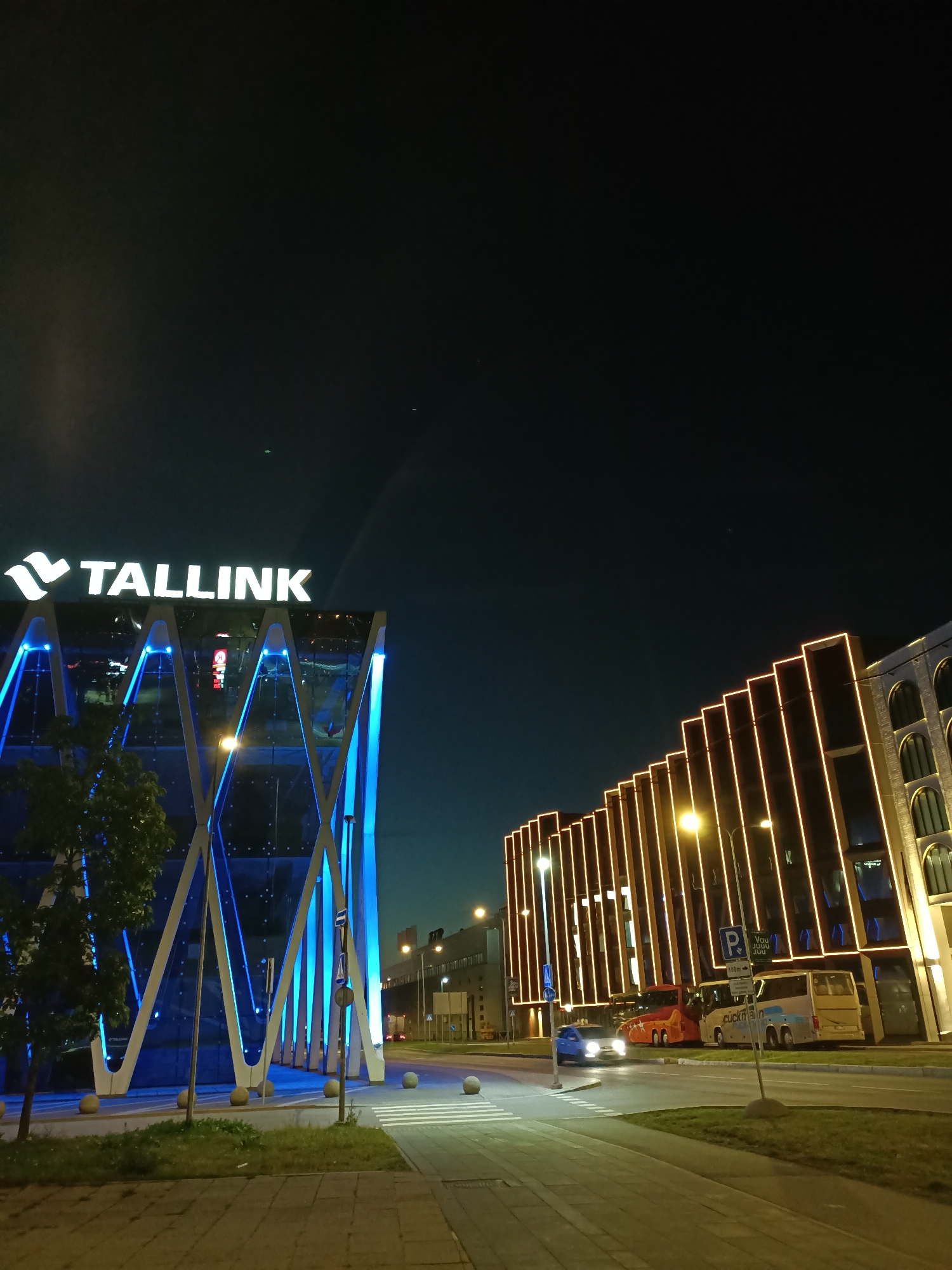 The image depicts a nighttime view of a street in Tallinn, Estonia, featuring modern architecture and illuminated signs. On the left, a glass building with the prominent "TALLINK" logo is illuminated by bright blue lights, highlighting its geometric facade. To the right, another building is adorned with vertical orange lights, creating a striking contrast. Several vehicles, including a blue car and two buses, are visible on the road. The scene is set against a dark night sky, with streetlights and building lights providing illumination. The overall impression is of a contemporary urban environment at night.