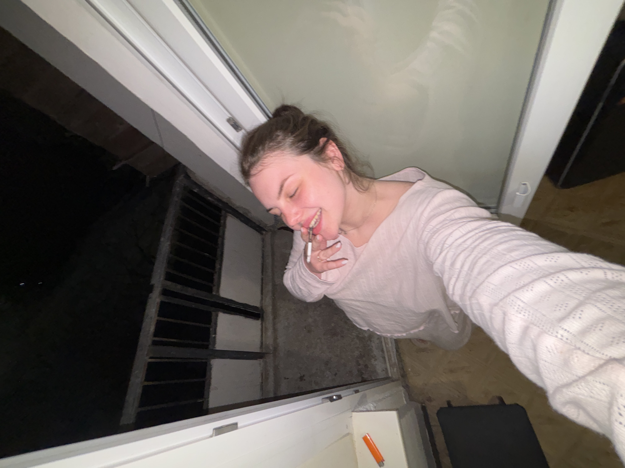 A woman is smoking a cigarette in an alcove. She is fair-skinned with brown hair pulled back in a bun, wearing a light-colored long-sleeved shirt. She has a ring on her right index finger. The alcove has a white-framed window or door, a metal grate, and a lighter on the sill. Beyond the grate, it is dark. The floor is brown and there is a dark-colored object near her feet. The image does not provide any indication that the location is Chornomorskoe, Ukraine.
