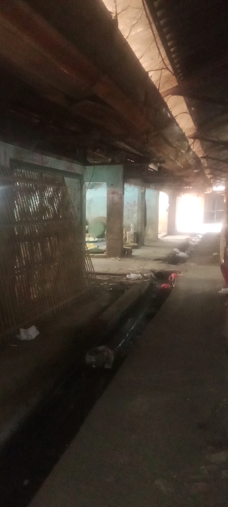 This is a photograph of a narrow, covered walkway or market stall in Karawang, Indonesia. The structure appears aged and in disrepair, with a corrugated metal roof supported by wooden beams, some of which are exposed and weathered. The walls are made of concrete or plaster, with some bearing graffiti. On the left side of the walkway, there is a large, woven bamboo or reed structure, possibly a partition or a display rack. The ground is concrete and appears dirty, with a dark, possibly water-filled, drainage channel running along the center of the walkway. Scattered trash, including plastic bags and what appear to be discarded items, is visible on the ground and in the channel. The lighting is dim and natural light is entering from the end of the walkway, creating a bright, overexposed area in the distance. No people are clearly visible in the photograph. The overall impression is one of a neglected or abandoned commercial space.