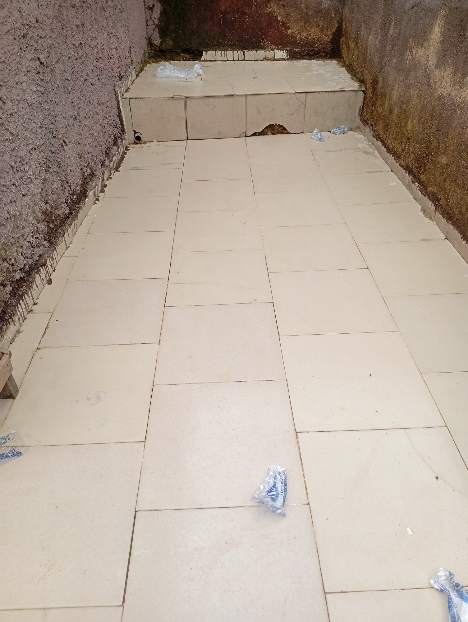 A tiled walkway is shown, leading from a set of tiered steps. The walls on either side are rough and textured, with evidence of water damage and staining. The tiled area has several pieces of clear plastic debris scattered on it. A small, gray pipe protrudes from the wall near the steps. The scene is outdoors, with the overall impression being one of disrepair and neglect. The location is identified as Rafin Par, Nigeria.