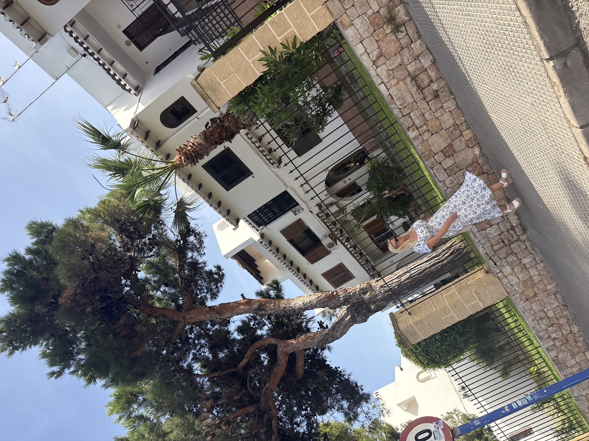 A woman in a blue and white floral dress and white wedges walks on a paved sidewalk beside a stone wall and a black metal fence. Behind the fence, there is a manicured lawn and a multi-story white building with dark shutters. A large tree with rough bark and dark green needles stands to the left of the woman, casting shadows on the sidewalk. In the background, more white buildings and a clear blue sky are visible. A circular speed limit sign is also visible on a blue pole in the lower left of the frame. The location is Urbanización Cabo Roig, Spain.
