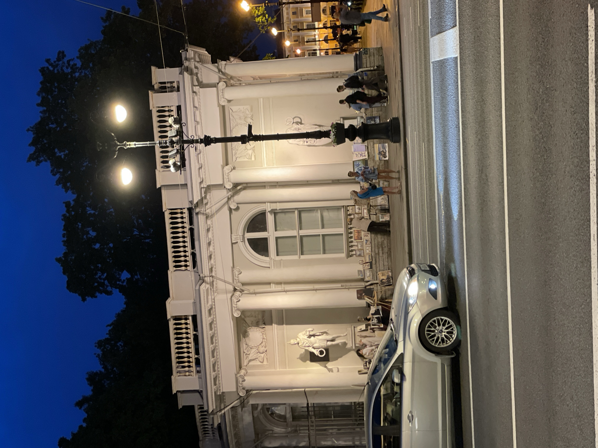 This image captures a street scene in Saint Petersburg, Russia, during twilight or early night, characterized by a dark blue sky and illuminated artificial lights. The prominent feature is a light-colored, classical-style building with a balustrade along its roofline and several large columns. A niche in the building houses a white statue of a classical male figure, possibly a Roman soldier, holding a wreath, illuminated from the left, casting a distinct shadow. Above the statue is a decorative relief sculpture.
On the sidewalk in front of the building, an open-air art market is active, with numerous paintings and artworks displayed on easels and leaned against the building wall. Several individuals are present: two women stand near the art displays, one in a blue dress; other people are seen further down the sidewalk, some browsing the art, others walking.
A silver Ford Focus sedan with its headlights and fog lights illuminated is in motion on the asphalt road in the foreground. A decorative black street lamp with two glowing, warm-toned lights stands prominently in the mid-ground, featuring a small flower planter at its base. Additional similar street lamps illuminate the street further into the distance. The road features white lane markings, and dark tree silhouettes are visible against the night sky behind the building, suggesting a nearby park or garden. The overall scene indicates the area around Arts Square (Ploshchad Iskusstv), known for its cultural institutions and outdoor art displays.