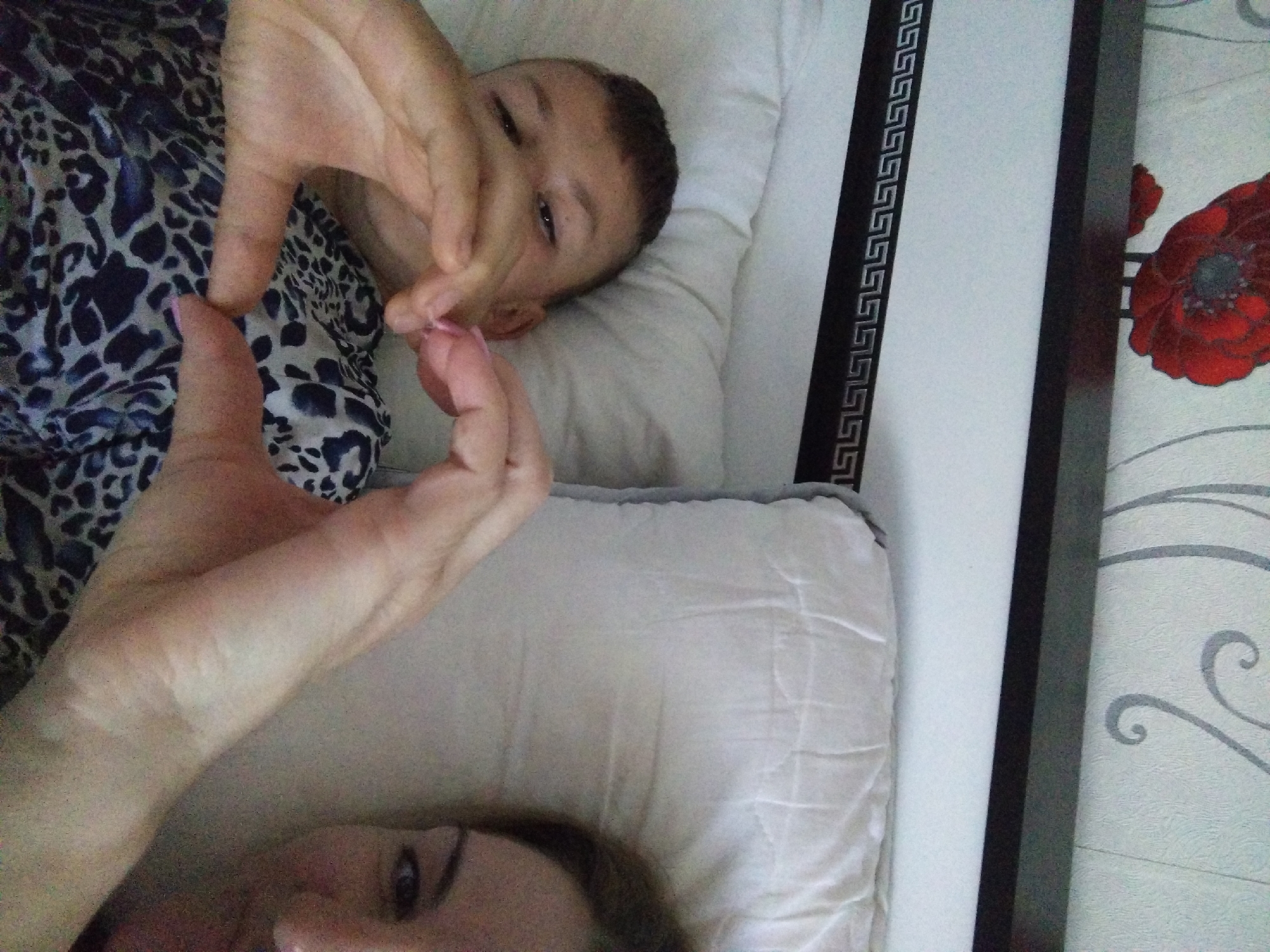 The image depicts a close-up, overhead view of two individuals lying down, forming a heart shape with their hands. A young person, likely a child or teenager, is positioned on the left side of the frame, with their face partially obscured by their hands. They have short, dark hair and are wearing a garment with a black and white leopard print. On the right side of the frame, the face of an adult, presumably female, is visible, with dark hair and visible eye makeup. She is also wearing a similar leopard print garment. The background consists of white pillows and a section of wallpaper featuring a red poppy design and grey swirling patterns. A dark wooden headboard with a Greek key border is also visible. The scene appears to be set in a bedroom. There is no information within the image to suggest its location in Stara Zagora, Bulgaria.