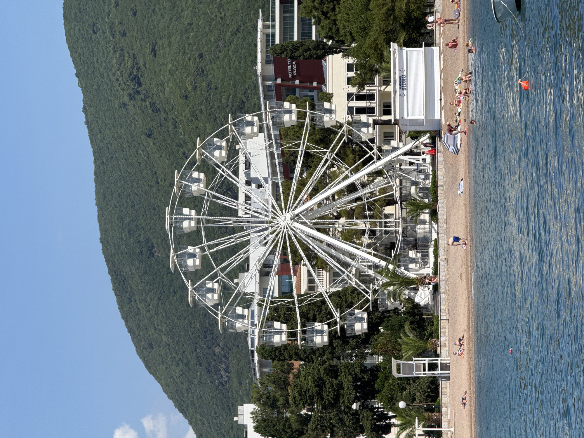 In Budva, Montenegro, a large white Ferris wheel stands prominently on a beachfront. The Ferris wheel is comprised of a circular structure with multiple enclosed cabins. Behind the Ferris wheel, a multi-story hotel with dark red accents is visible, along with other buildings and lush green trees. The foreground features a sandy beach with several people sunbathing and a few individuals in the water. A white kiosk with the word "SIDRO" on it is also present on the beach. The sea is calm and blue, and a small boat is docked to the right of the frame. A steep, tree-covered mountain rises in the background under a clear blue sky.