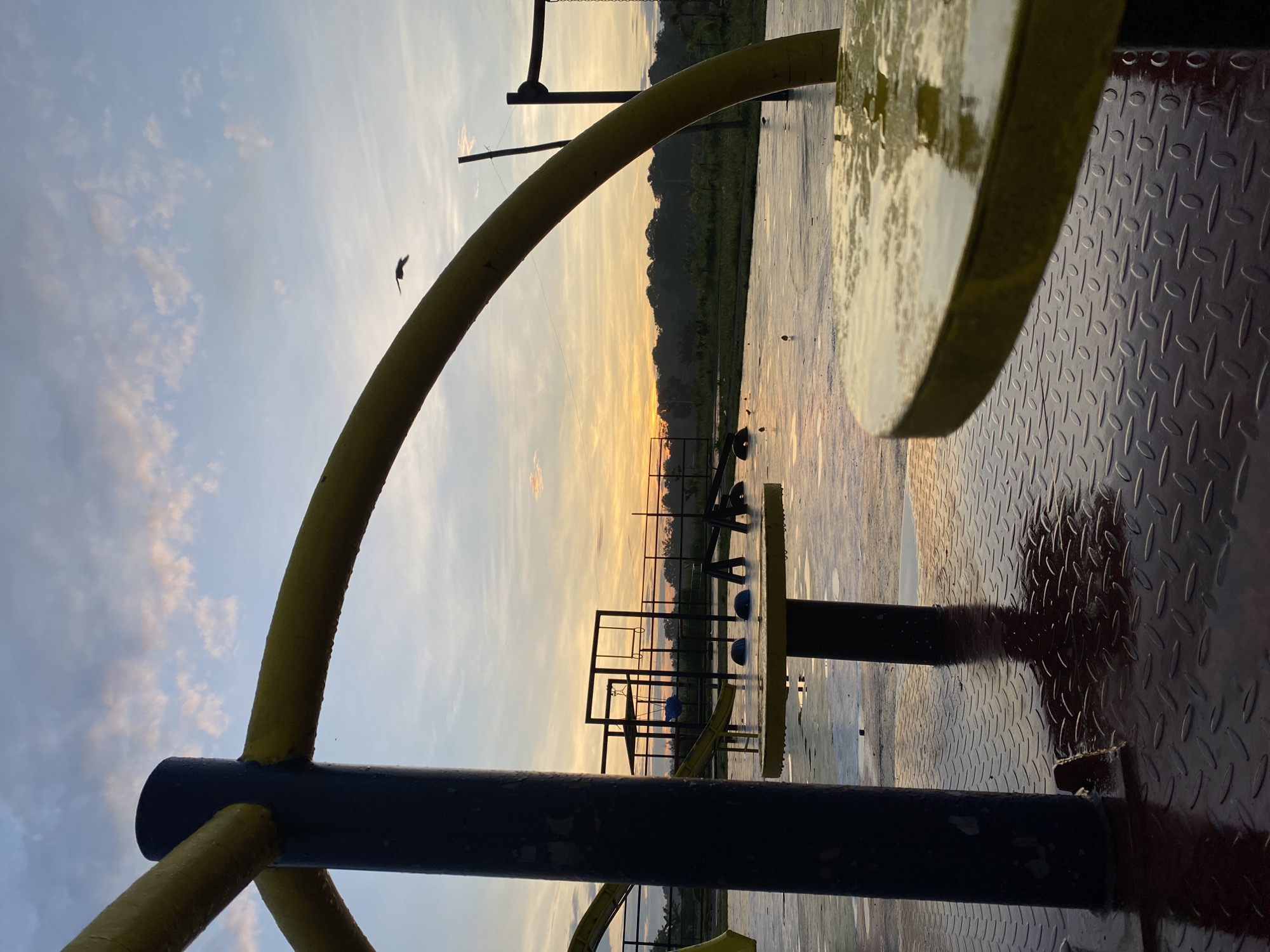 The image captures a serene sunset scene over a body of water, likely in Davao City, Philippines. The perspective is taken from a platform or deck, looking out towards the horizon.
In the foreground, a prominent structure of black and yellow metal pipes or bars extends across the frame. A large, curved yellow bar arches from the bottom left towards the upper right. Part of a wet, yellow, reflective circular surface is visible on the far right, reflecting the ambient light. Below this, a textured red diamond plate surface, possibly a deck or walkway, is visible and appears wet.
The midground features an expansive body of calm water, which beautifully reflects the sky's colors, transitioning from light blue to warm orange and yellow. Silhouetted against the glowing horizon are several dark metal structures, possibly a pier, dock, or elements of a water park, some appearing partially submerged. A few dark, rounded objects are also floating in the water.
In the background, a dark, undulating line of land, likely forested hills or a distant shoreline, stretches along the horizon. The sky above is a canvas of soft blues with scattered, wispy clouds, blending into vibrant hues of orange and yellow where the sun is setting. A small bird is captured mid-flight against the open sky in the upper left portion of the image.
The scene conveys a tranquil atmosphere, with the warm light of the sunset illuminating the wet surfaces and reflecting off the water. No individuals or visible text are present in the image.