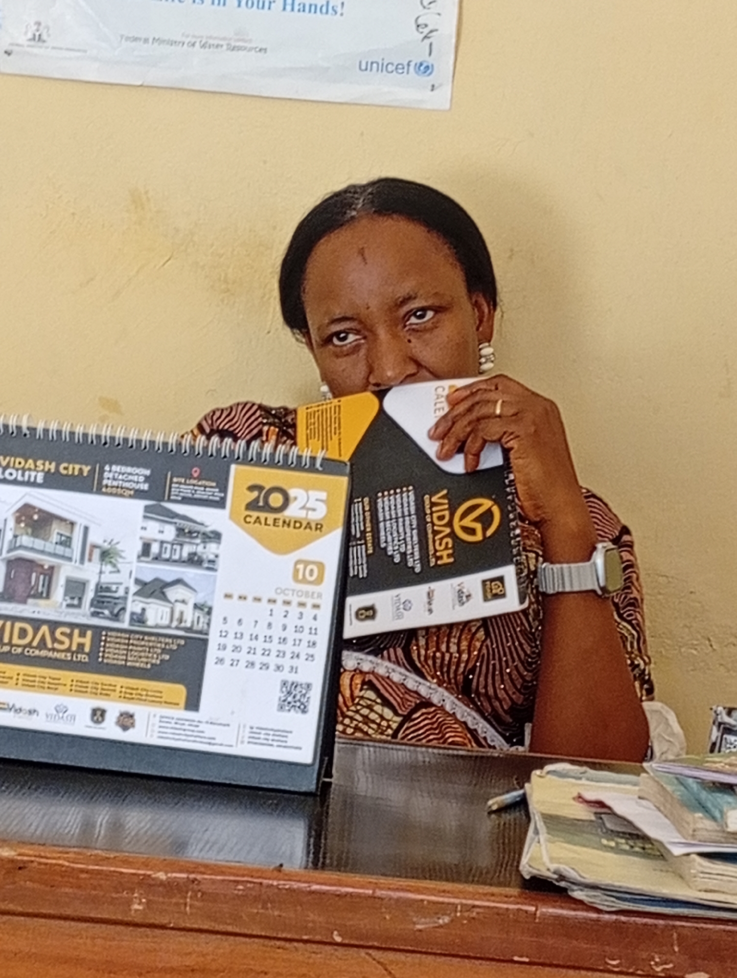 A woman is seated at a desk, partially obscuring her face with a 2025 calendar and a black booklet. The calendar is branded "Vidash City" and shows a month view for October with the 10th circled. Text on the calendar includes references to "4 Bedroom Detached Penthouse" and lists "Vidash Group of Companies Ltd" and "Vidash City Shelters Ltd," and the phrase "Our own dreams Estate". The booklet is also branded "Vidash" and partially hides the woman's face. She wears a patterned brown top, earrings, a wedding band on her ring finger, and a watch on her left wrist. The desk is cluttered with papers and documents. A poster with the word "Unicef" is visible on the wall in the background. The calendar also mentions Narayi.